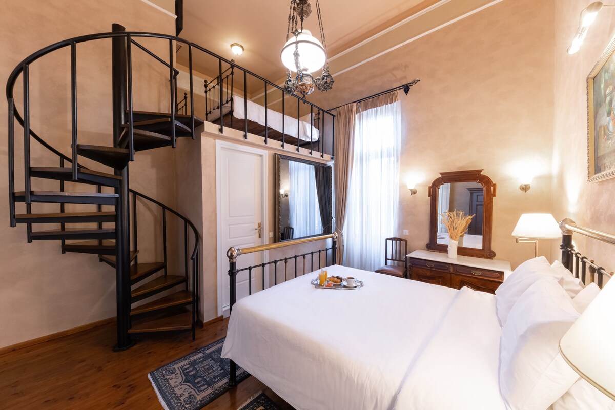Un dormitorio con cama, candelabro, escalera en espiral, cómoda, espejo y ventana con cortinas.