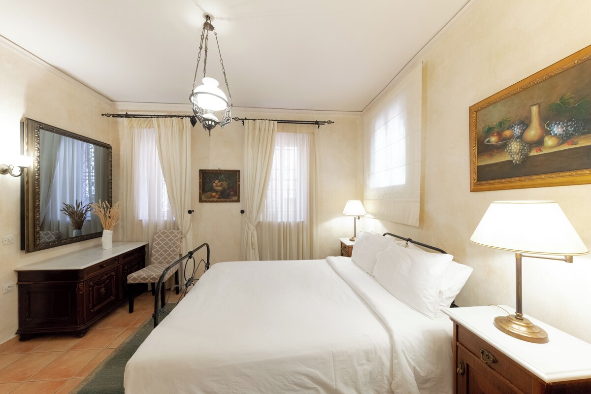 Habitación de hotel con una cama grande, un escritorio con silla, cómoda, espejo, un cuadro y una lámpara.