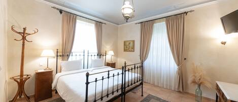 Un dormitorio con una cama con dosel, dos mesitas de noche con lámparas, un espejo grande, un televisor montado en la pared y una ventana con cortinas.