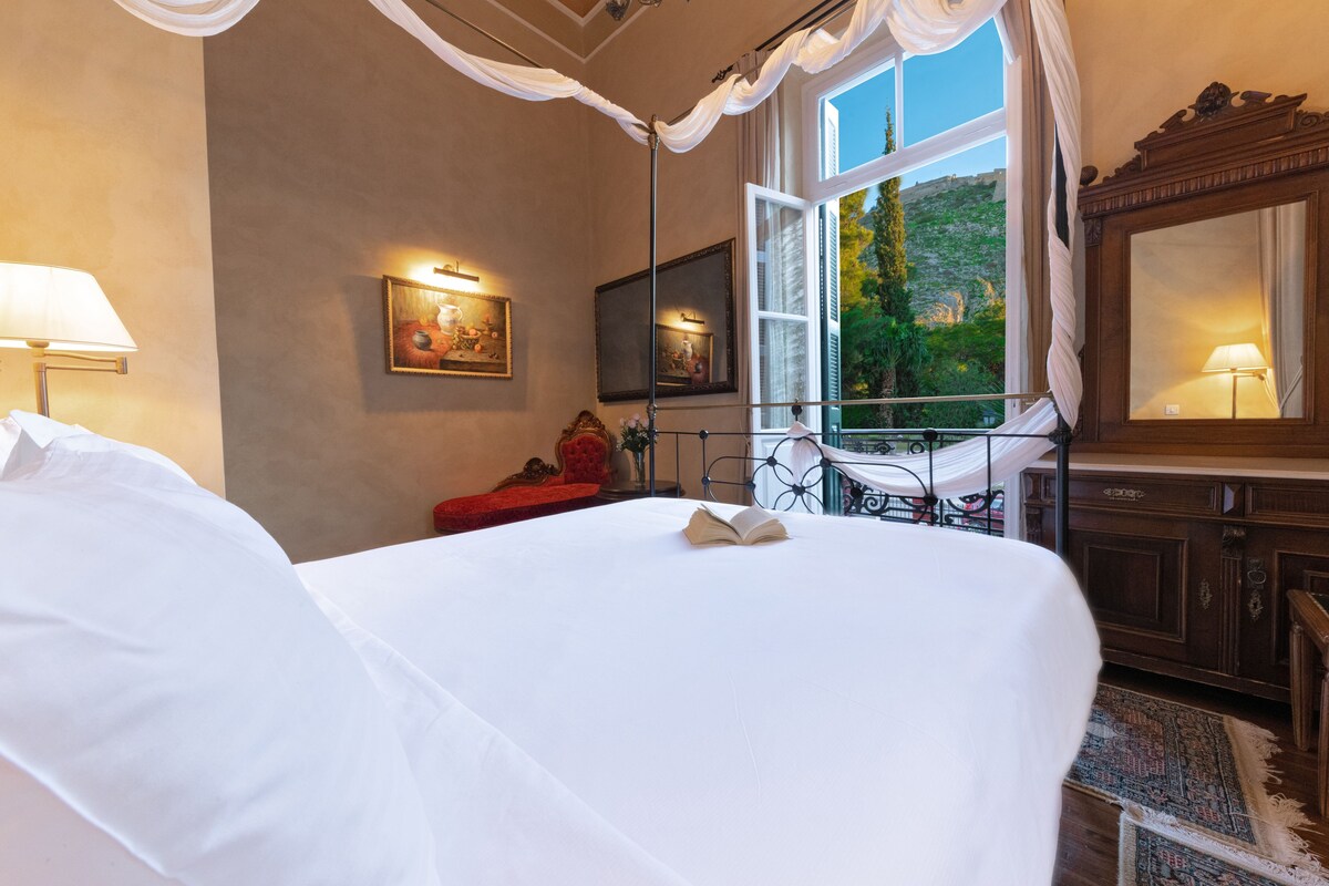 Habitación de hotel con una cama grande, ventana con vistas a vegetación, un espejo y una silla roja.
