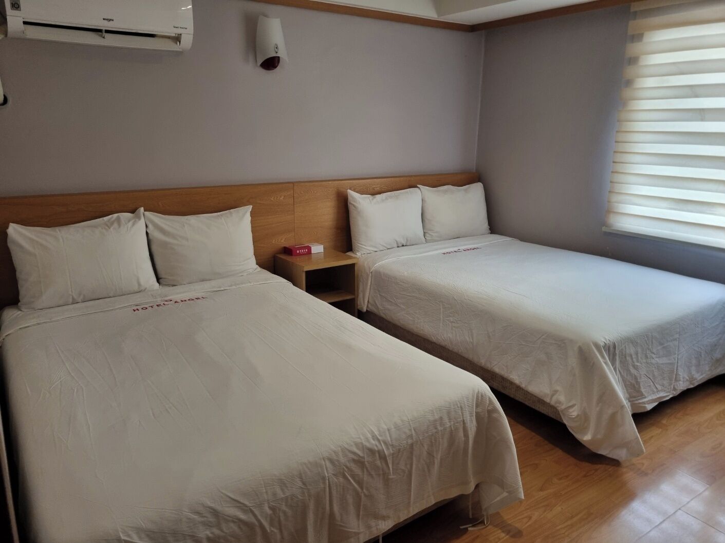 A hotel room with two single beds, a wooden headboard, a bedside table with a red object, and a window with blinds.