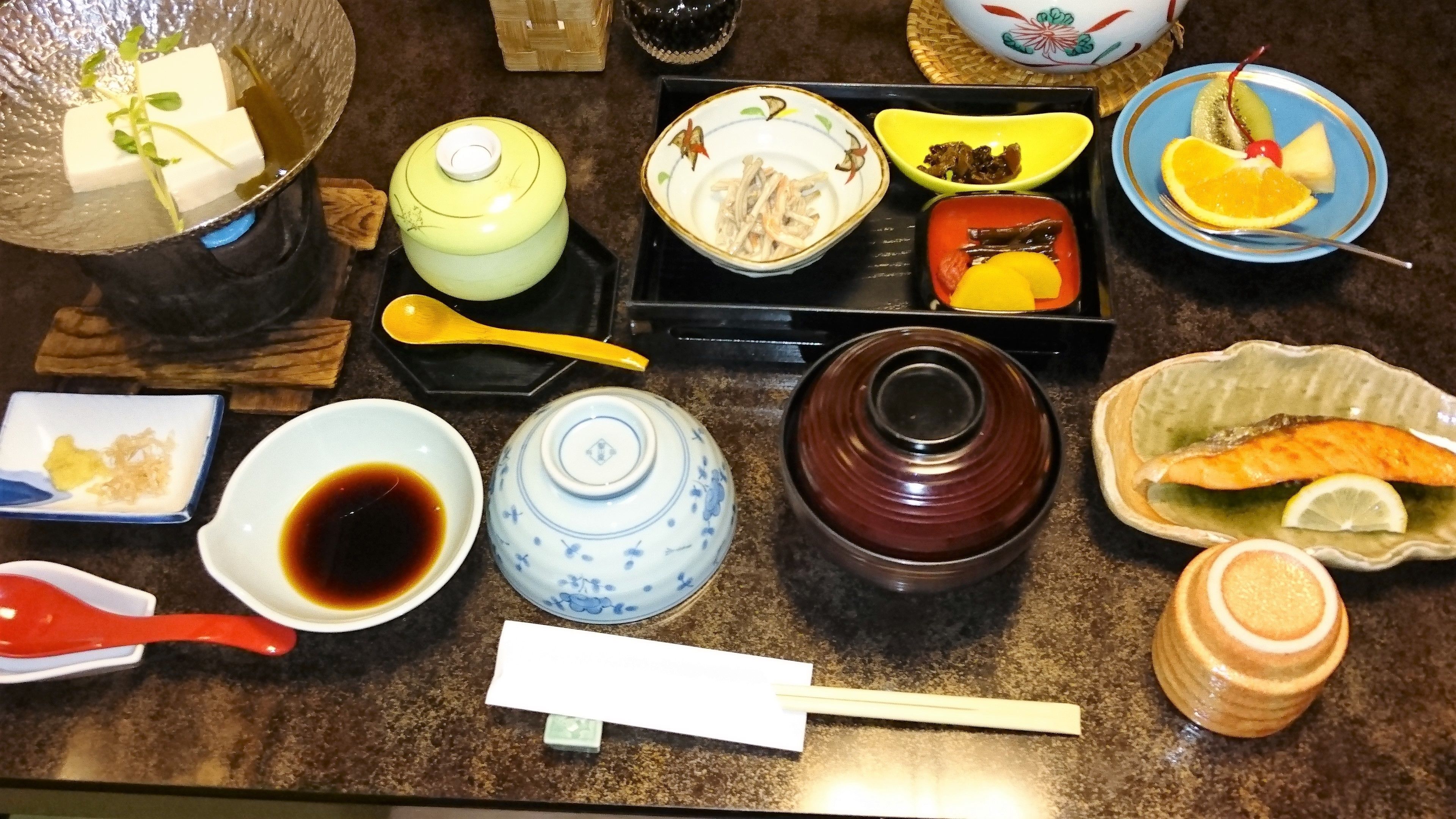 A variety of Japanese dishes and utensils on a table, including a green pot, a blue and white teapot, a brown ceramic container, and a blue plate with a fish and lemon slice.
