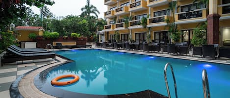 A swimming pool with lounge chairs and a lifebuoy, surrounded by a building with balconies and potted plants.