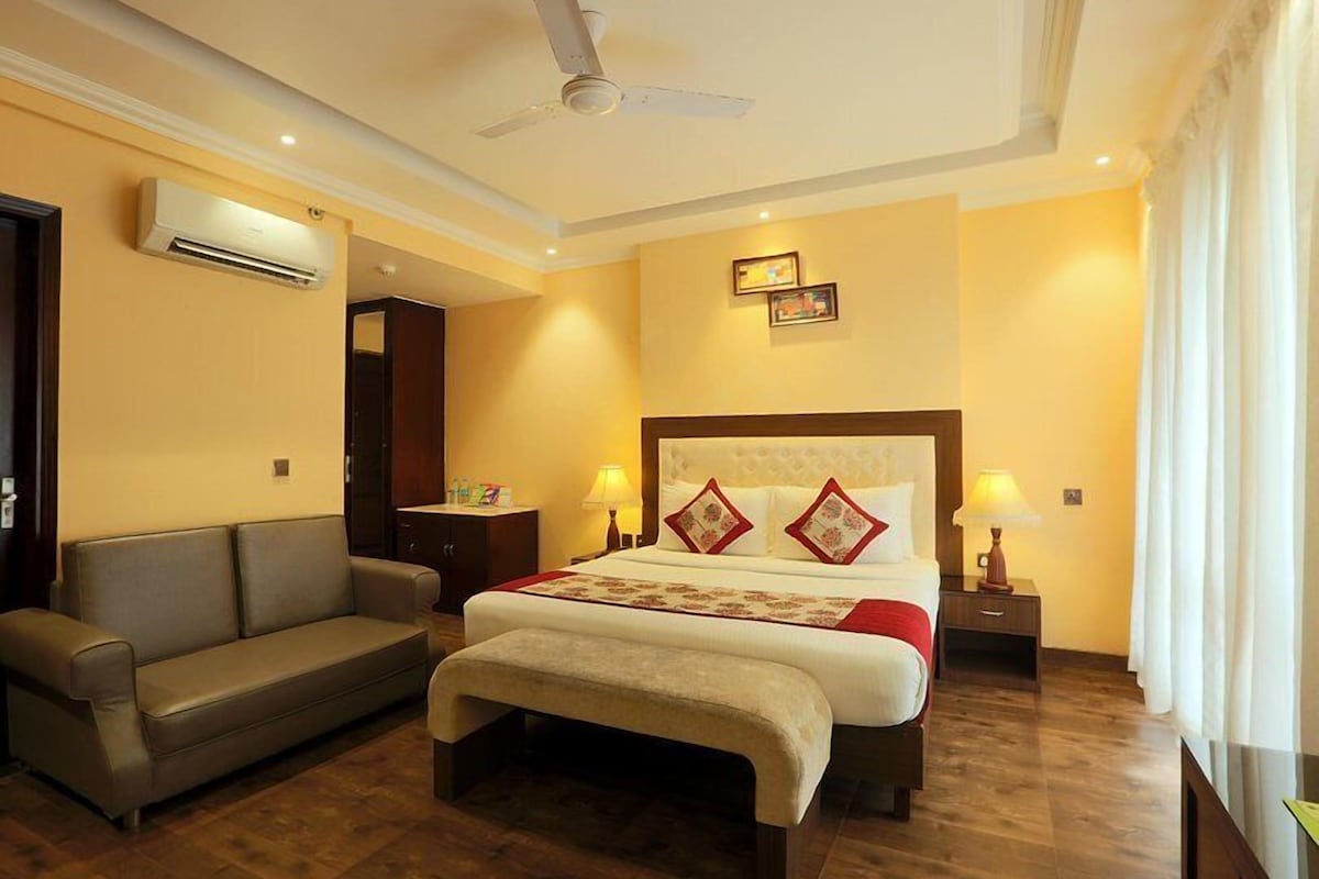 A hotel room with a bed, a sofa, a ceiling fan, an air conditioning unit, and two bedside lamps.
