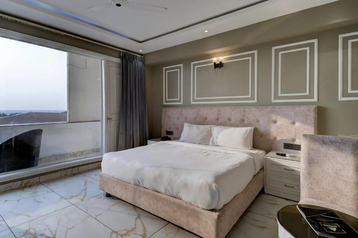 A modern hotel room with a large bed, a bedside table, a chair, and a balcony with a view.