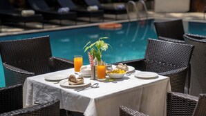 A dining table set with plates, cutlery, glasses, and a bowl of food, surrounded by wicker chairs, with a pool in the background. - De Alturas Resort (Candolim)