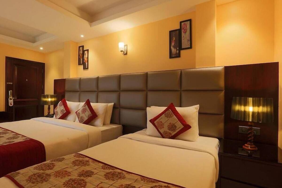 A hotel room with two beds, a wooden headboard, a nightstand with a lamp, and a wall-mounted light fixture.