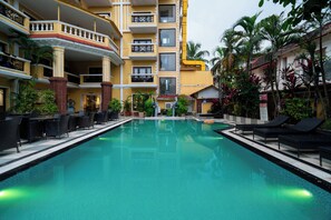 Ein Pool mit Liegestühlen und Tischen, umgeben von einem mehrstöckigen Gebäude mit Balkonen und tropischen Pflanzen.