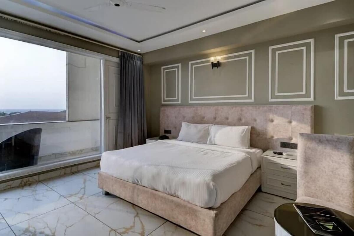 A modern hotel room with a large bed, a bedside table, a chair, and a balcony with a view.