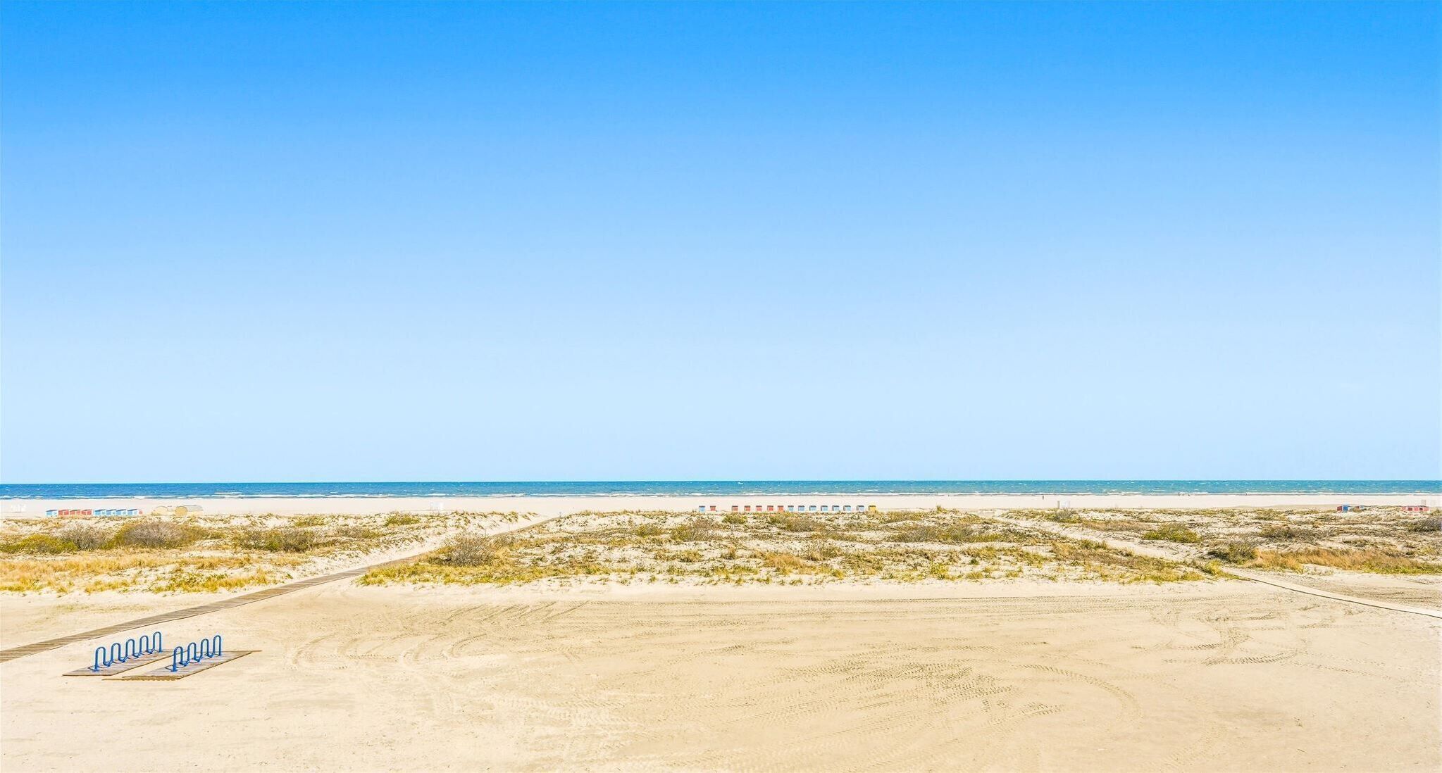 Une plage avec un ciel dégagé, un sol sablonneux et quelques bâtiments visibles au loin.