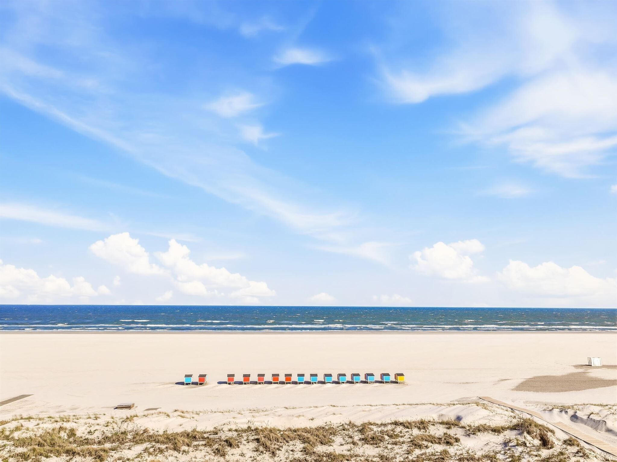 Une plage bordée d’une rangée de cabanes colorées et surplombée d’un ciel bleu clair.