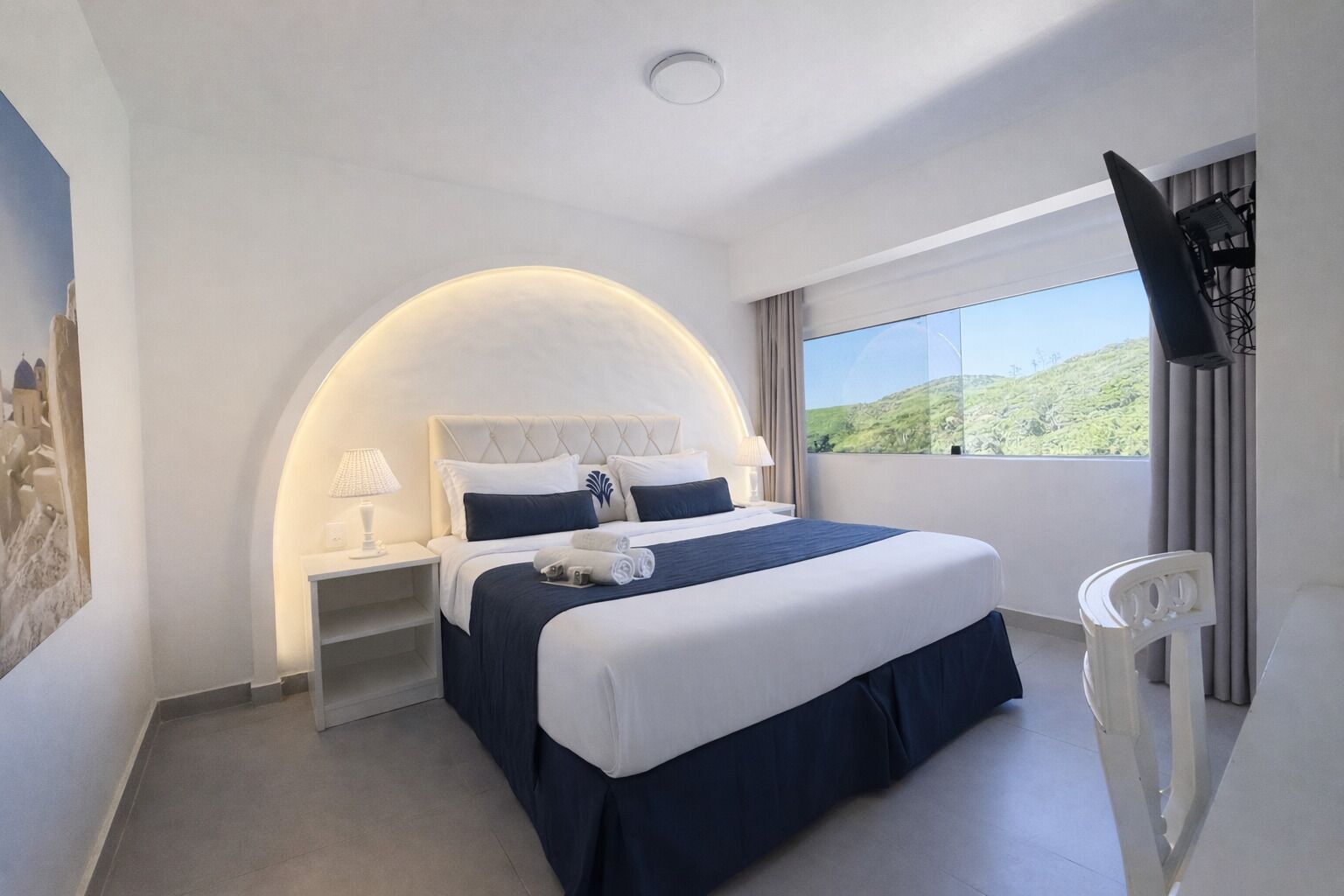 Un cuarto de hotel con cama, mesita de noche, televisor, ventana con vistas y un cuadro en la pared.