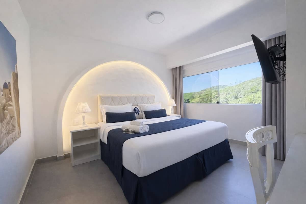 Un cuarto de hotel con cama, mesita de noche, televisor, ventana con vistas y un cuadro en la pared.