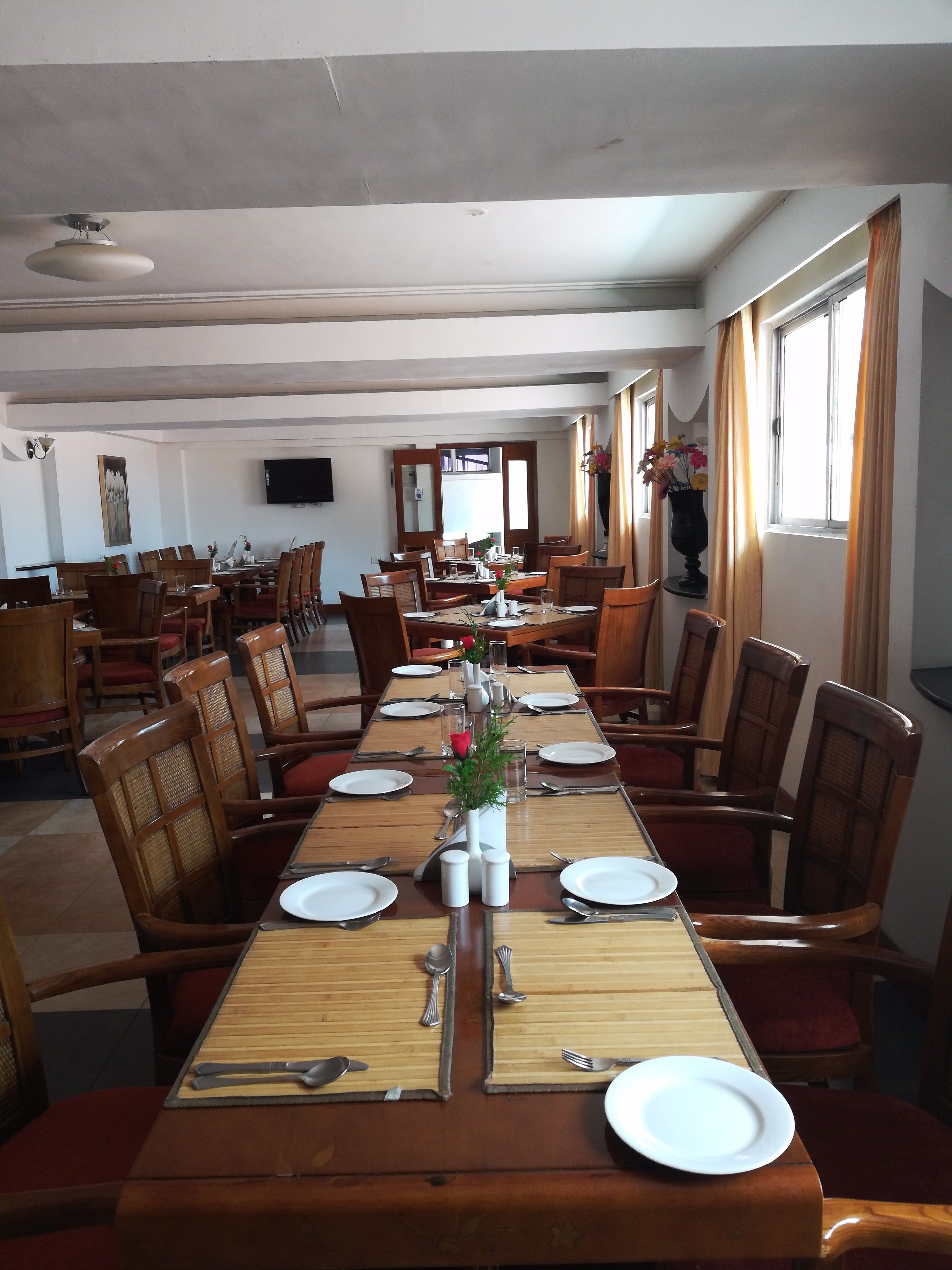 A dining area with multiple tables set for meals, wooden chairs, a television mounted on the wall, and a window with curtains.