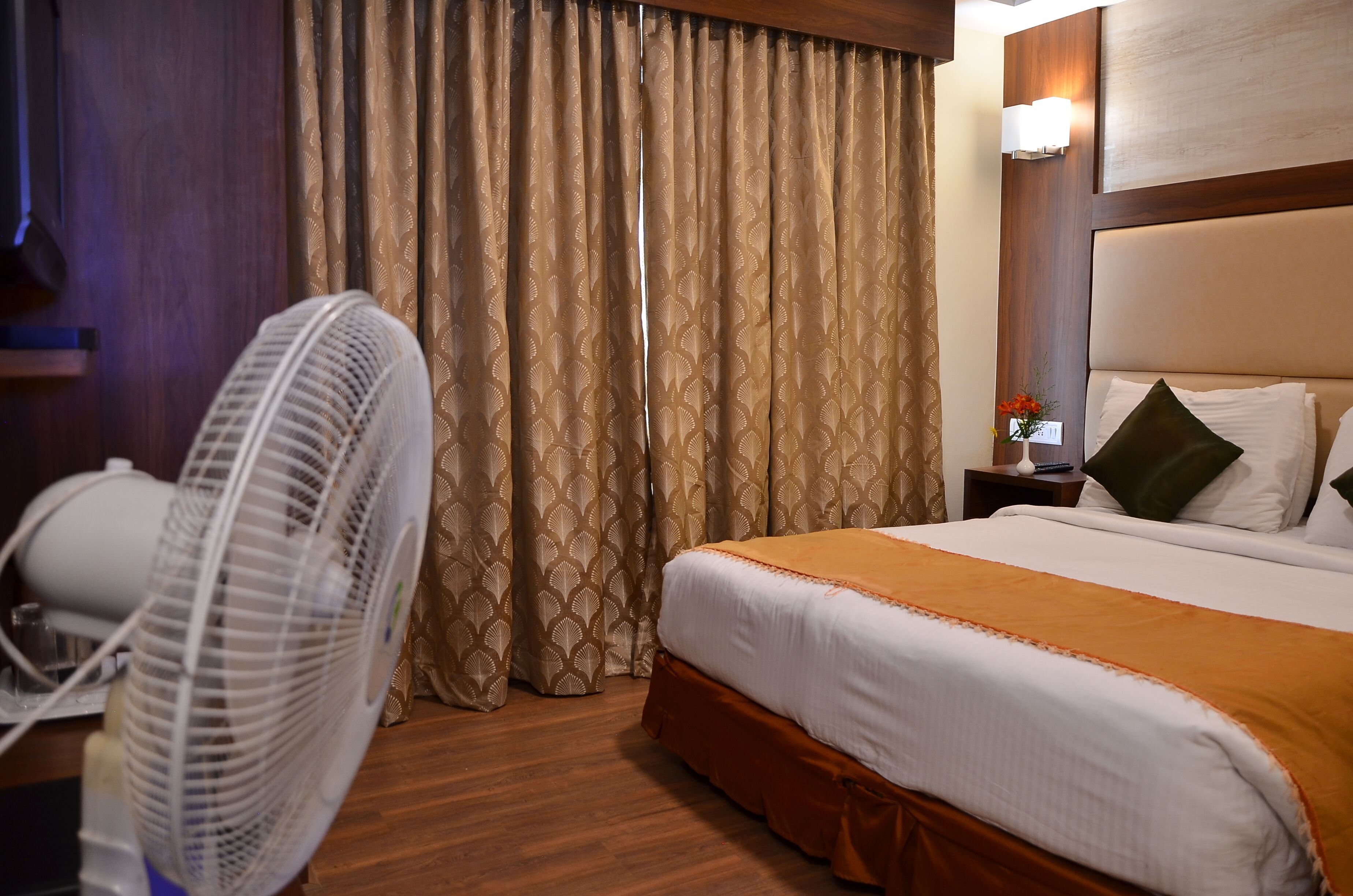 A hotel room with a bed, a standing fan, and a nightstand with a lamp and a vase.