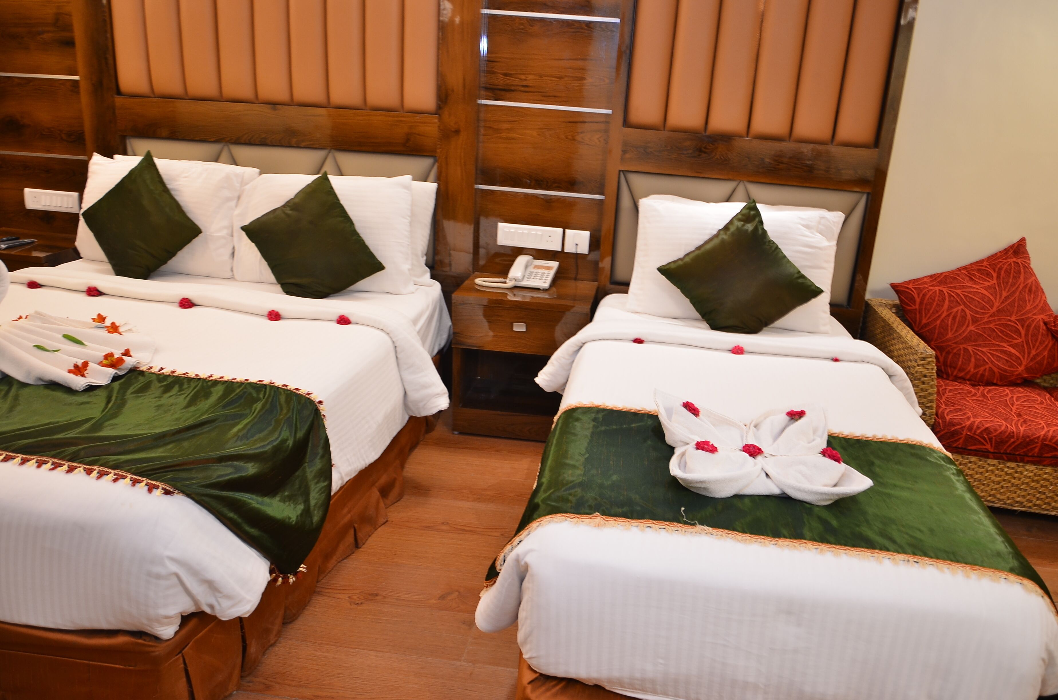 A hotel room with two beds, each with green and white bedding, a wooden headboard, a bedside table with a phone, and a red patterned pillow.