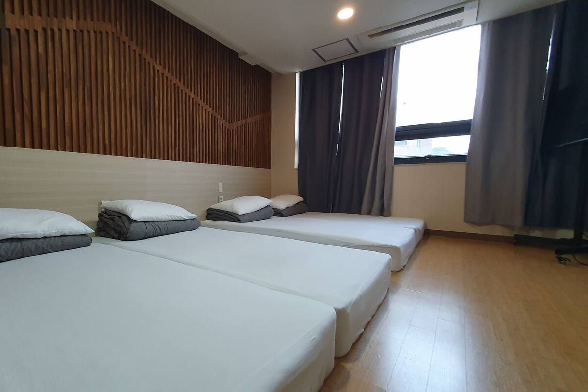 Habitación de hotel con tres camas, paneles de madera en la pared, una ventana con cortinas y un equipo de aire acondicionado.
