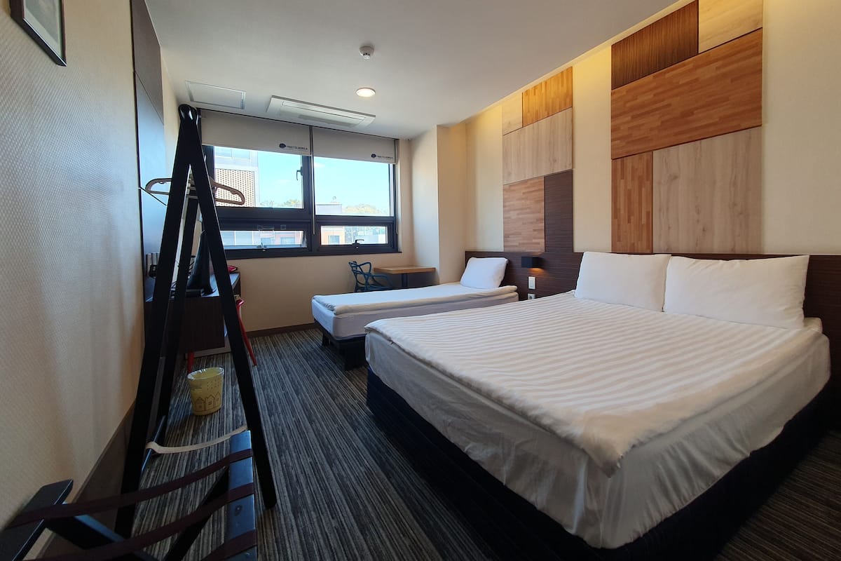 Habitación de hotel con dos camas, un escritorio, una silla y una ventana con vistas a edificios.