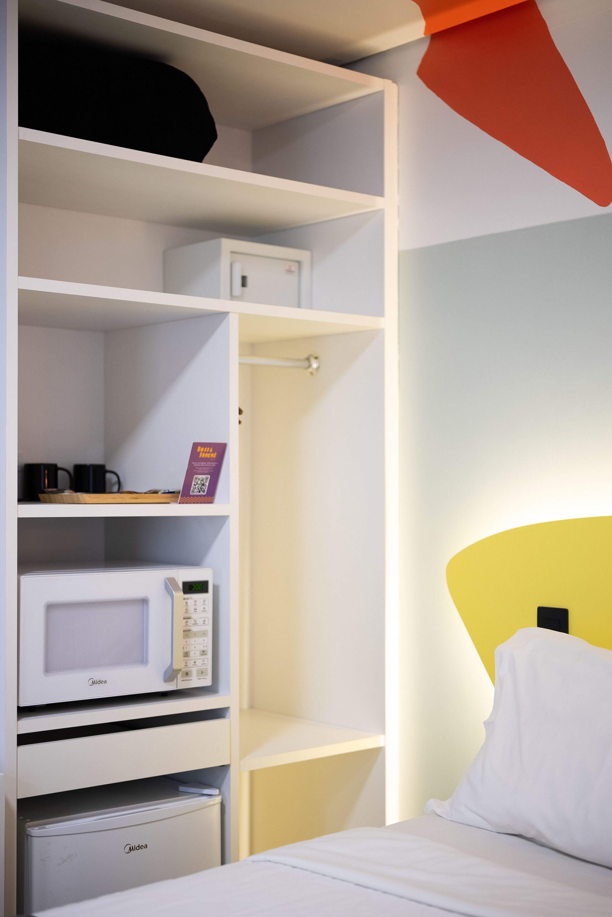 Habitación de hotel con cama blanca, microondas blanco, mini refrigerador y estante con cafetera y un cartel con código QR.