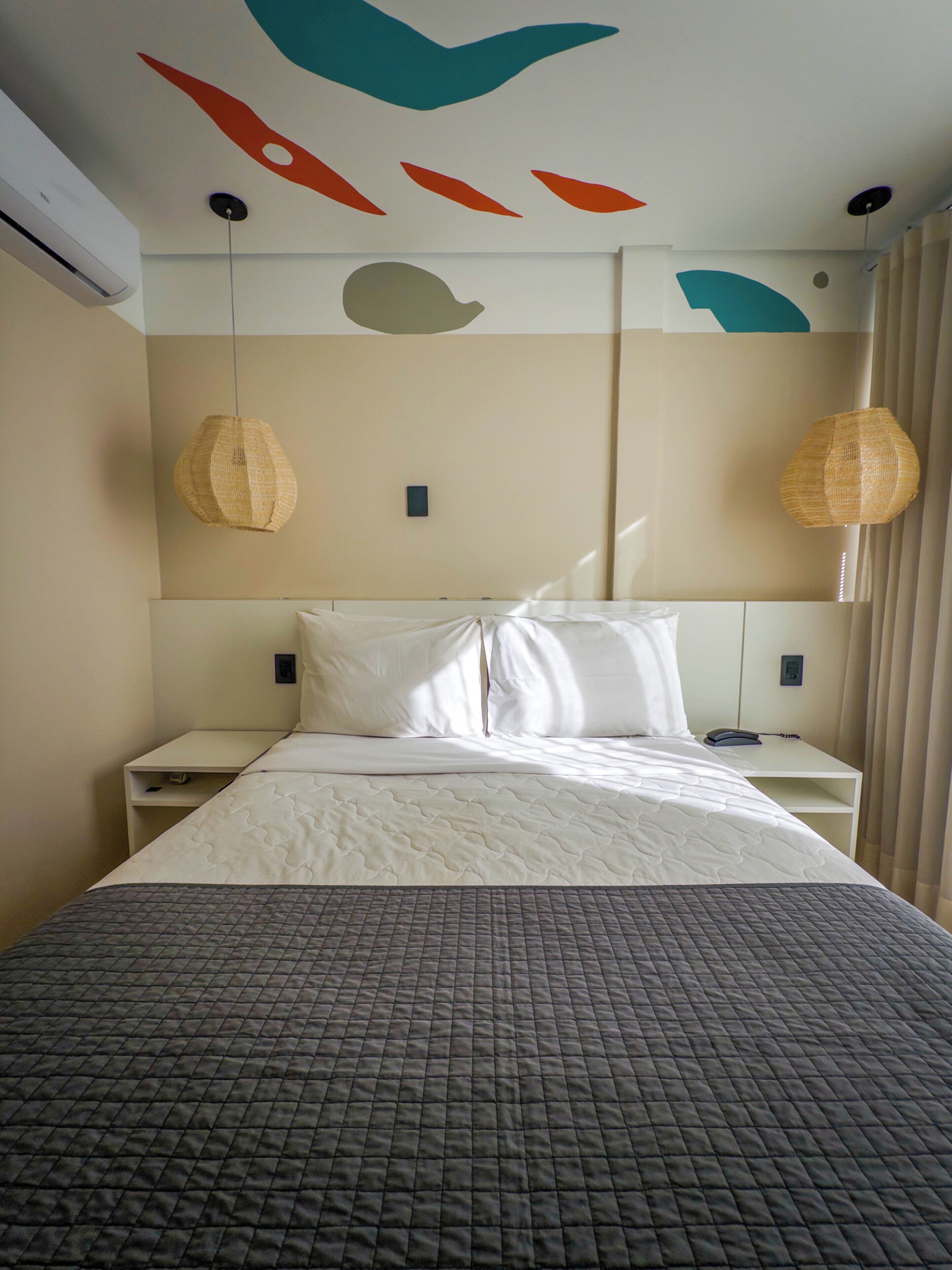 Une chambre d’hôtel comprenant un lit, deux tables de chevet, un climatiseur fixé au mur et un plafond orné d’œuvres d’art murales abstraites.