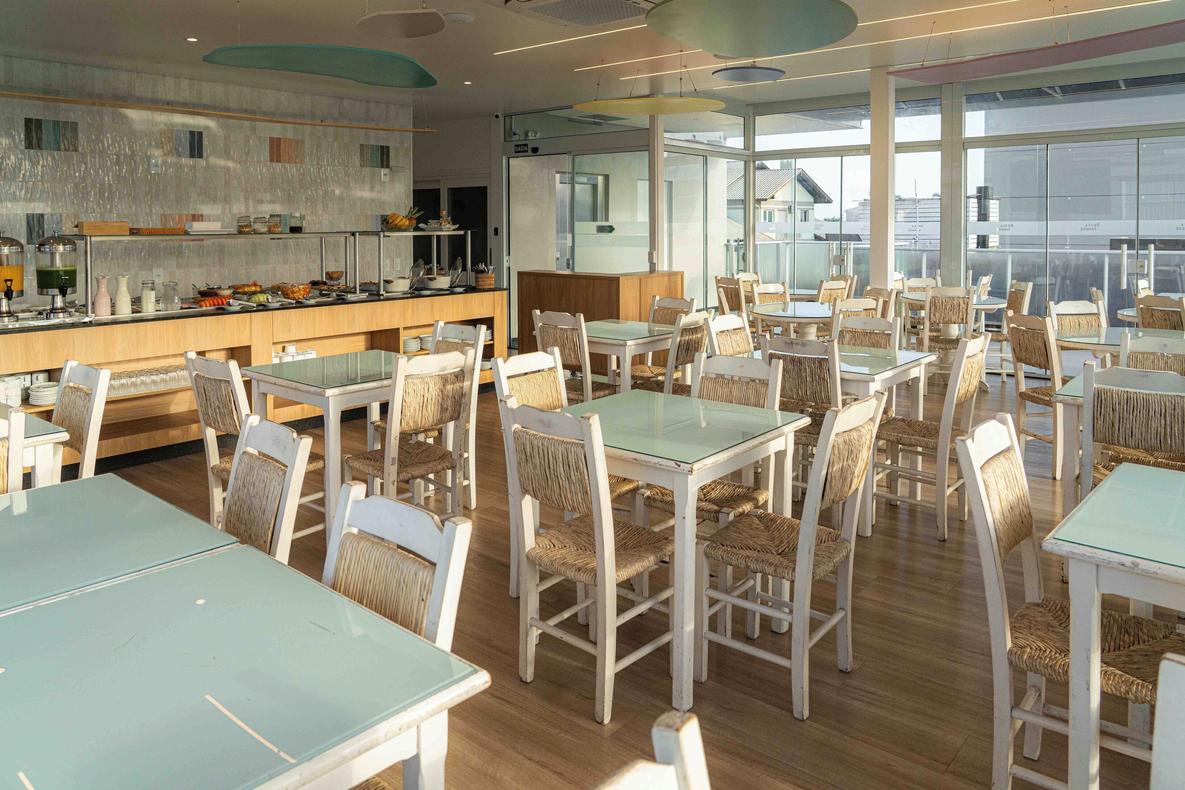 A spacious dining area with multiple tables and chairs, a counter with food items, and large windows providing natural light.