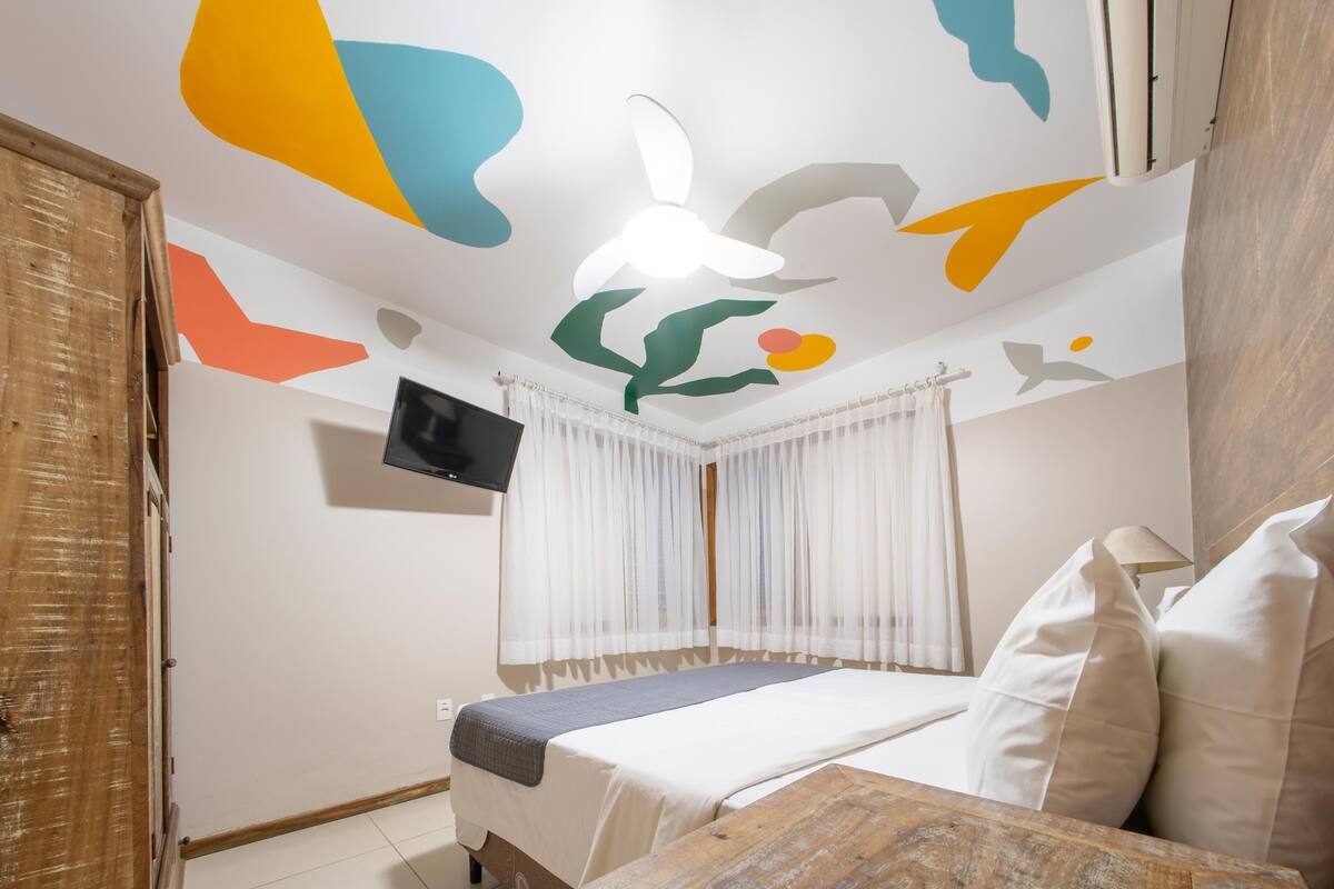A bedroom with a bed, a flat-screen TV, a ceiling fan, and a window with sheer curtains.