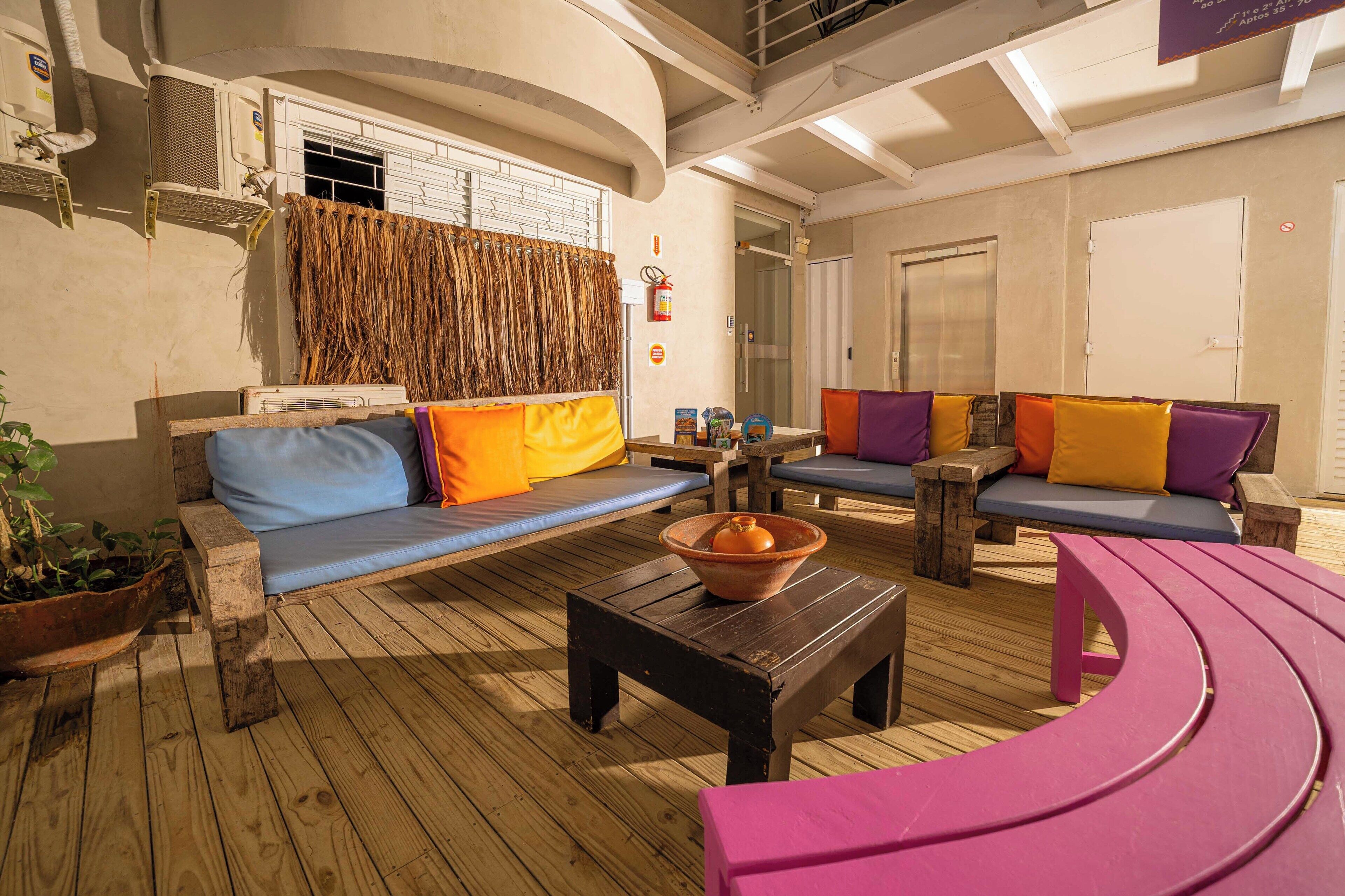 Un espacio interior bien iluminado con asientos, piso de madera, sofás acolchados con cojines de colores, un cuenco de terracota sobre una mesa de madera y una planta en maceta.