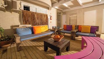 A sunlit indoor seating area with wooden flooring, cushioned sofas with colorful cushions, a terracotta bowl on a wooden table, and a potted plant.
