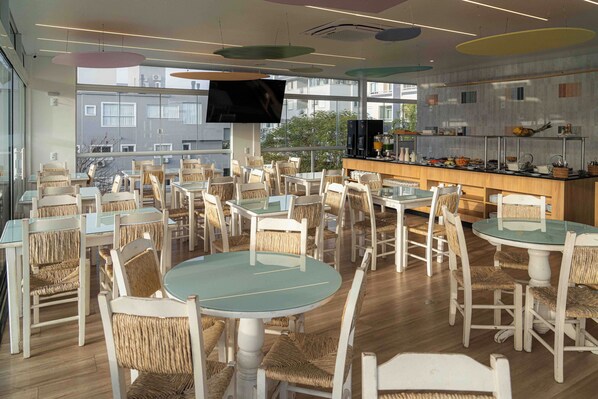 Un moderno comedor con mesas redondas, sillas de mimbre, una pantalla de televisión grande y un mostrador de buffet con diferentes opciones de comida.