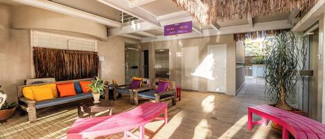 A sunlit lounge area with wooden flooring, a thatched ceiling, and a sofa with colorful cushions.