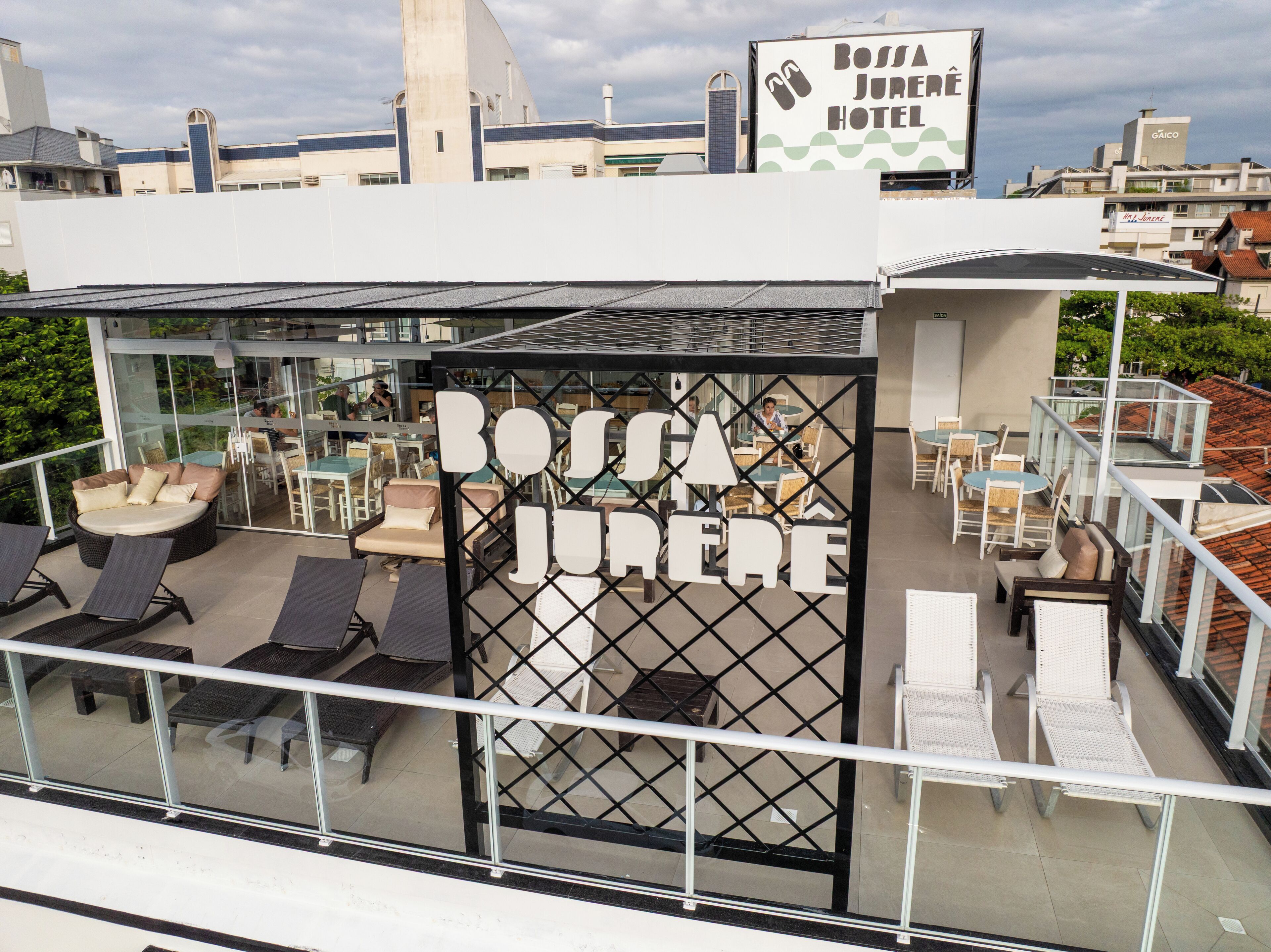 A rooftop lounge area with outdoor seating and a glass enclosure displaying the name "Bossa Jurê Hotel".