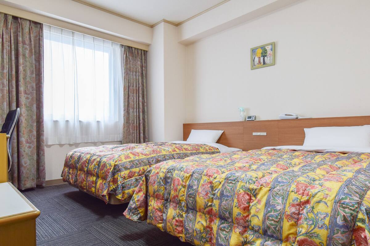 Quarto de hotel com duas camas, cabeceira de madeira, uma mesa pequena, uma cadeira, janela com cortinas e um quadro na parede.