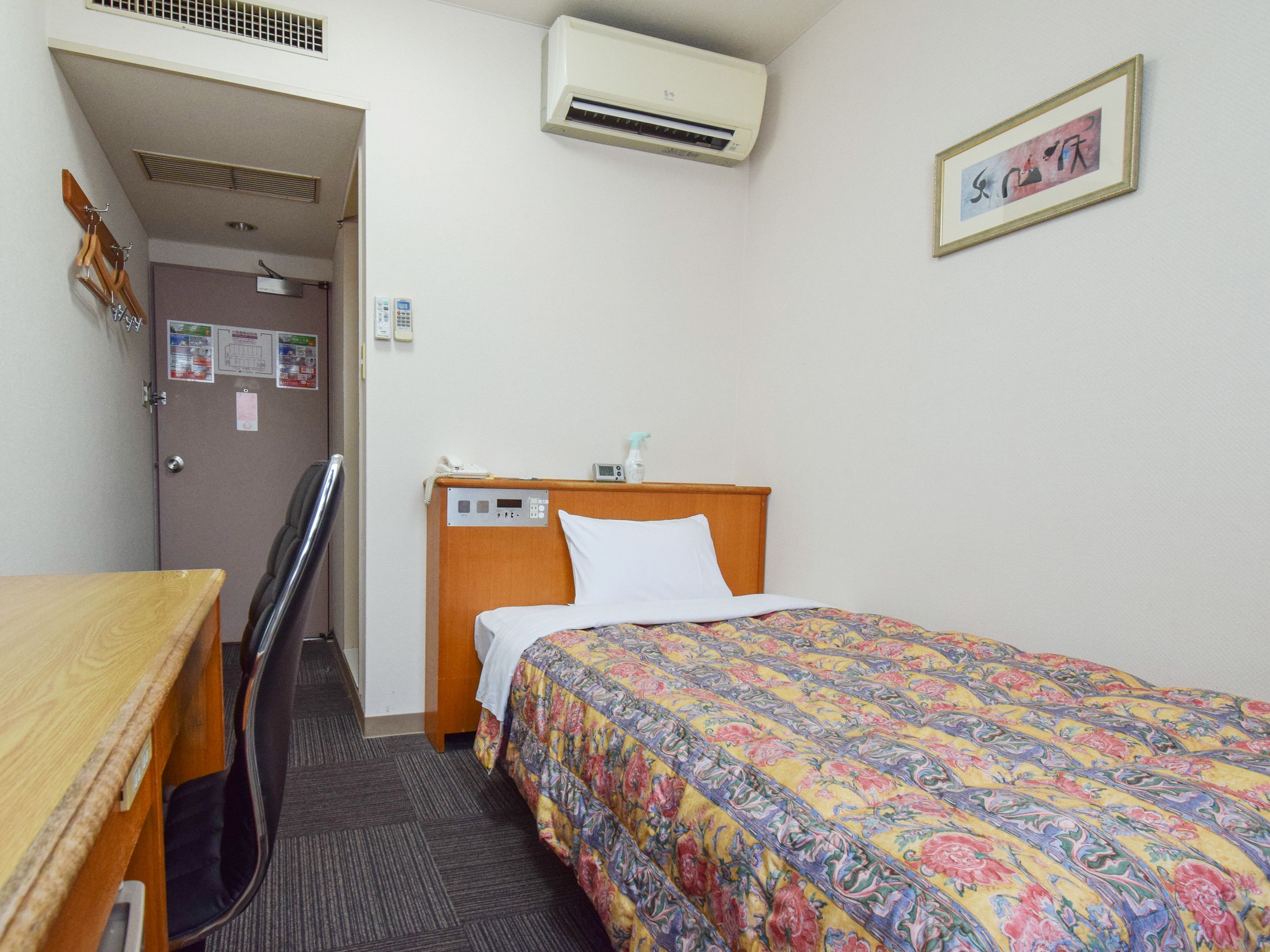 Une chambre d’hôtel avec un bureau en bois, un lit recouvert d’une courtepointe à motifs floraux colorés, une chaise, un climatiseur et une porte ornée d’une affiche.