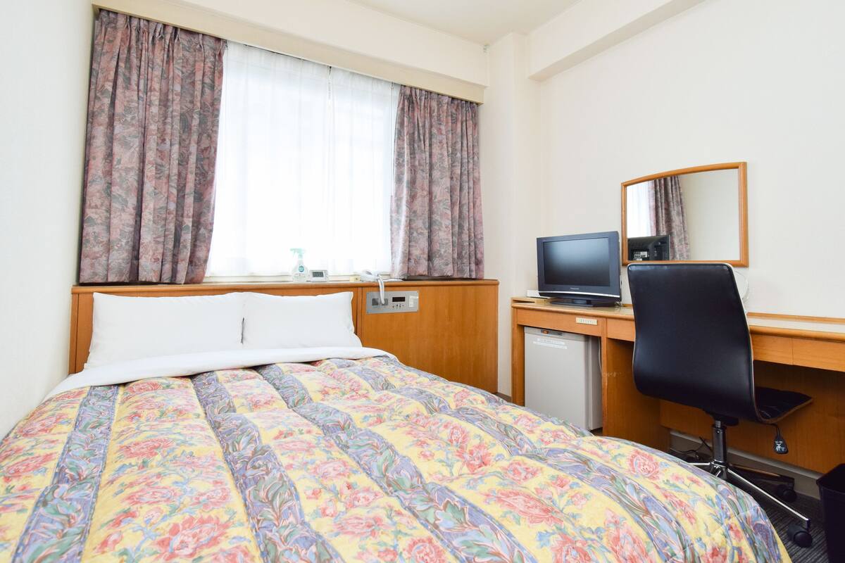 Quarto de hotel com cama, escrivaninha com computador, cadeira, pequeno frigorífico e janela com cortinas.