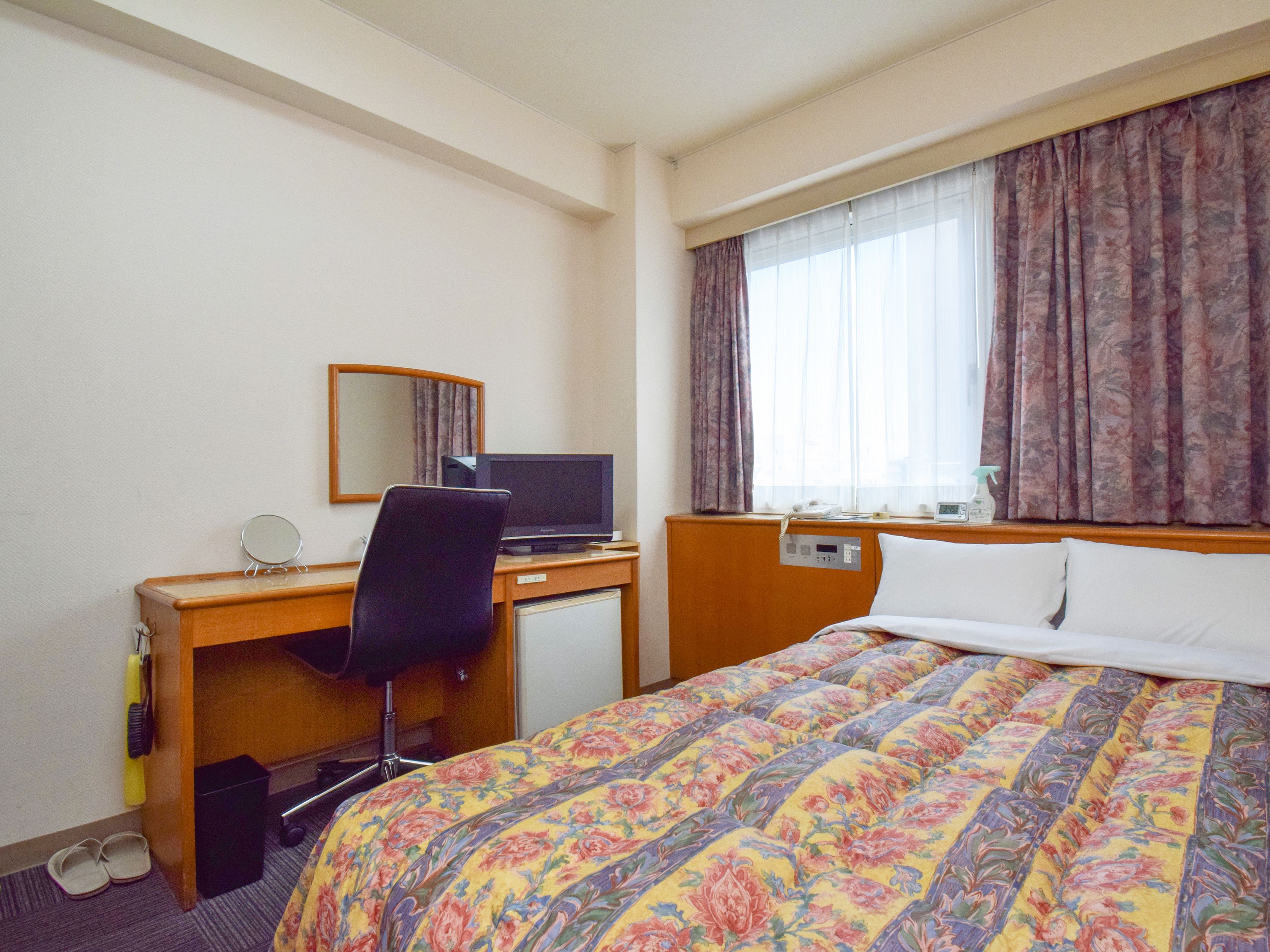 A hotel room with a bed, a desk with a computer, a chair, a small table, a mirror, and a window with curtains.