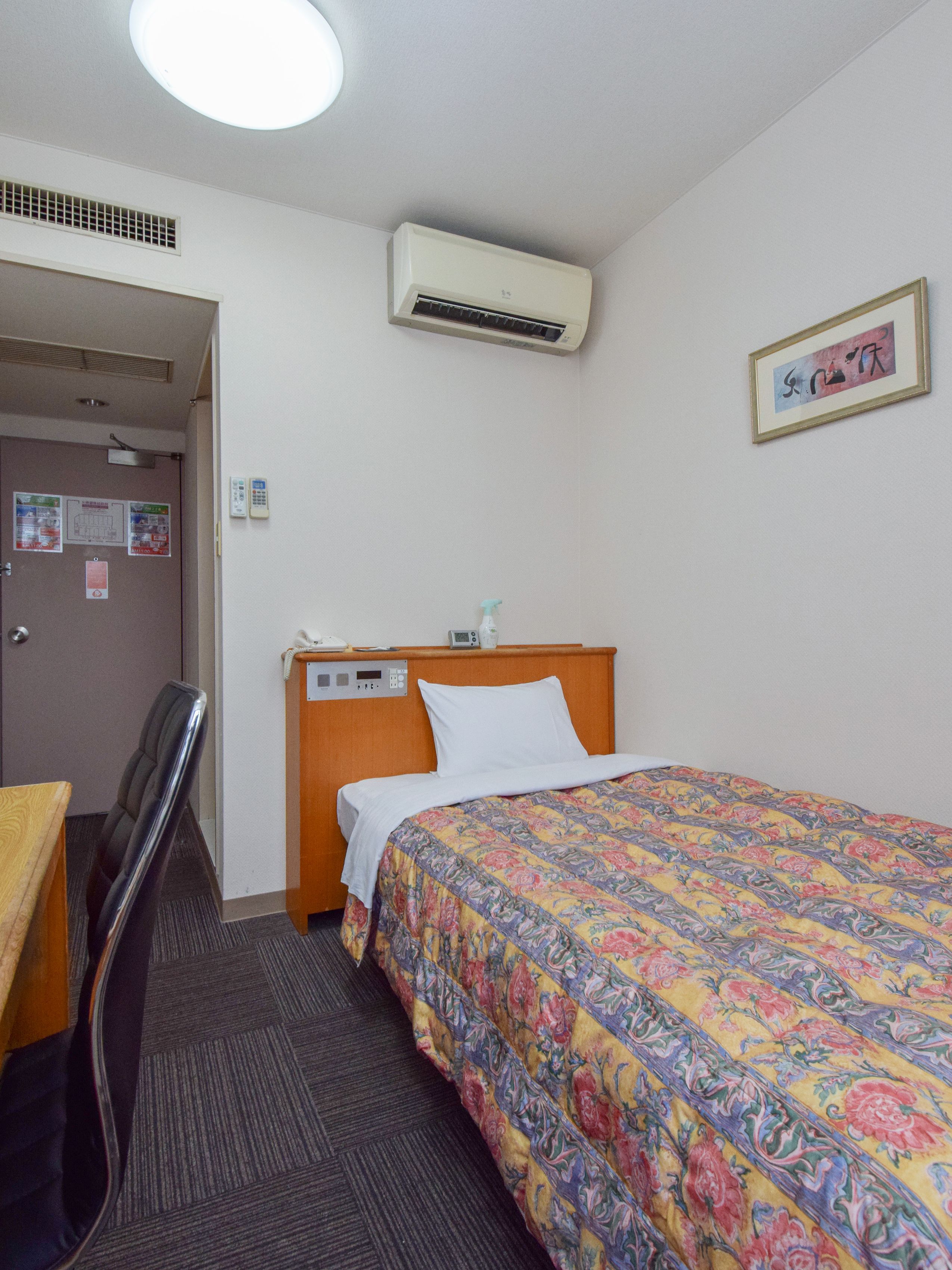 Ett hotellrum med en säng, ett skrivbord, en stol, en luftkonditionering och en inramad tavla på väggen.