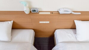 Een hotelkamer met twee witte bedden, een nachtkastje met een flesje, een digitale klok, een telefoon en een stopcontact.