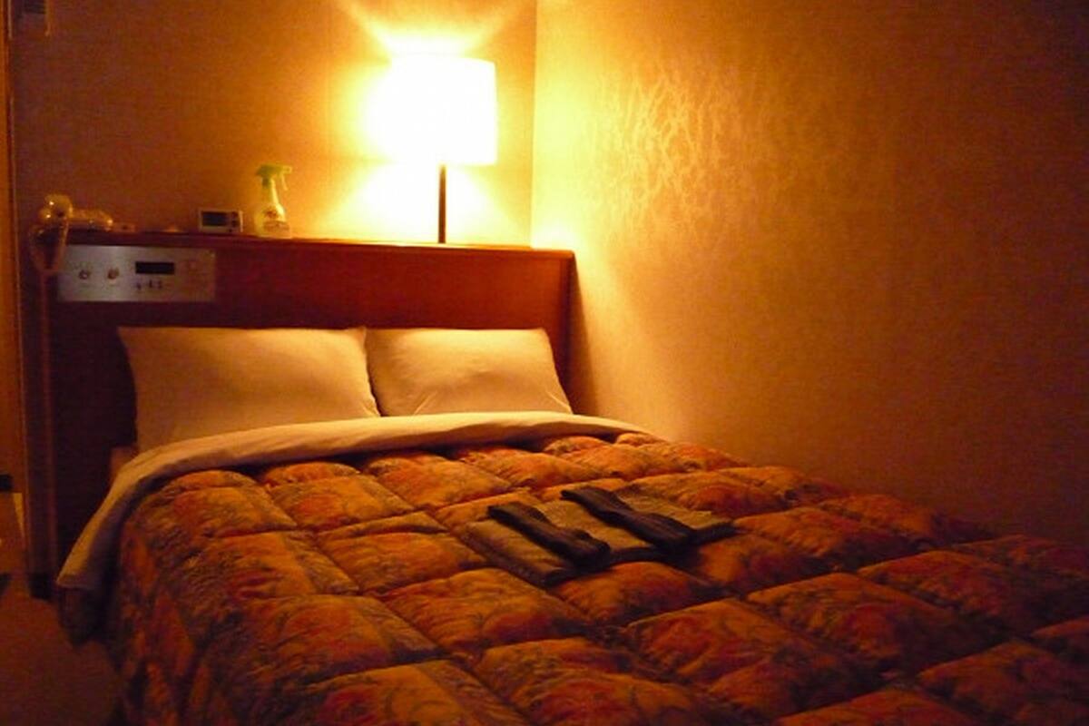 Quarto de hotel com cama, mesa de cabeceira, abajur e relógio.