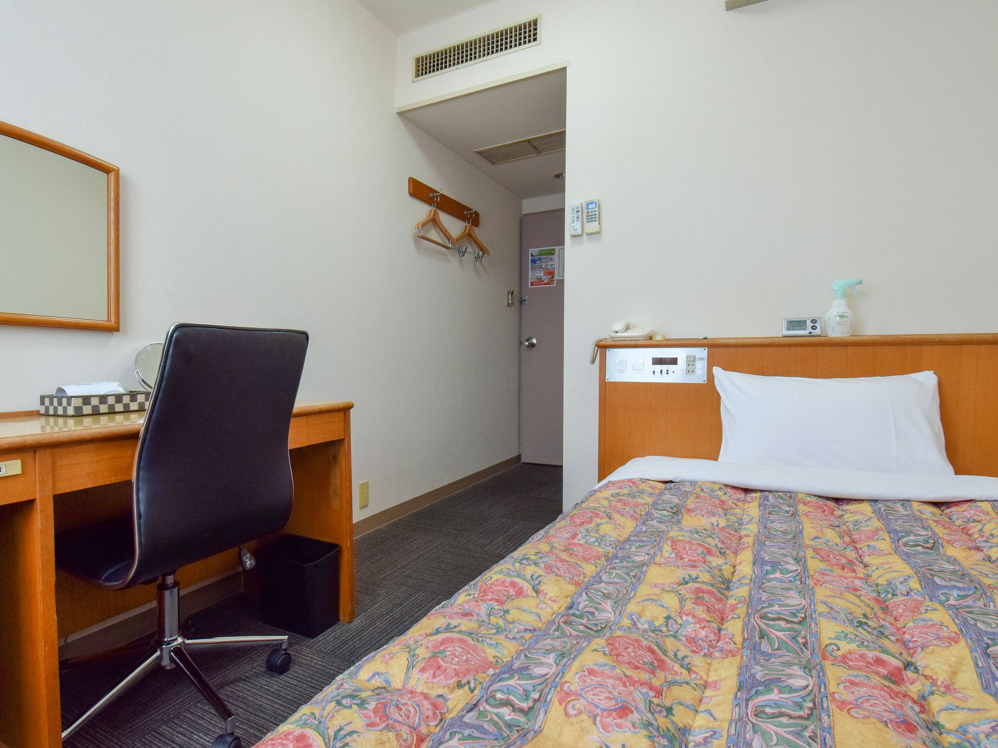 A hotel room with a bed, a desk, a chair, a mirror, a telephone, and a small clock.