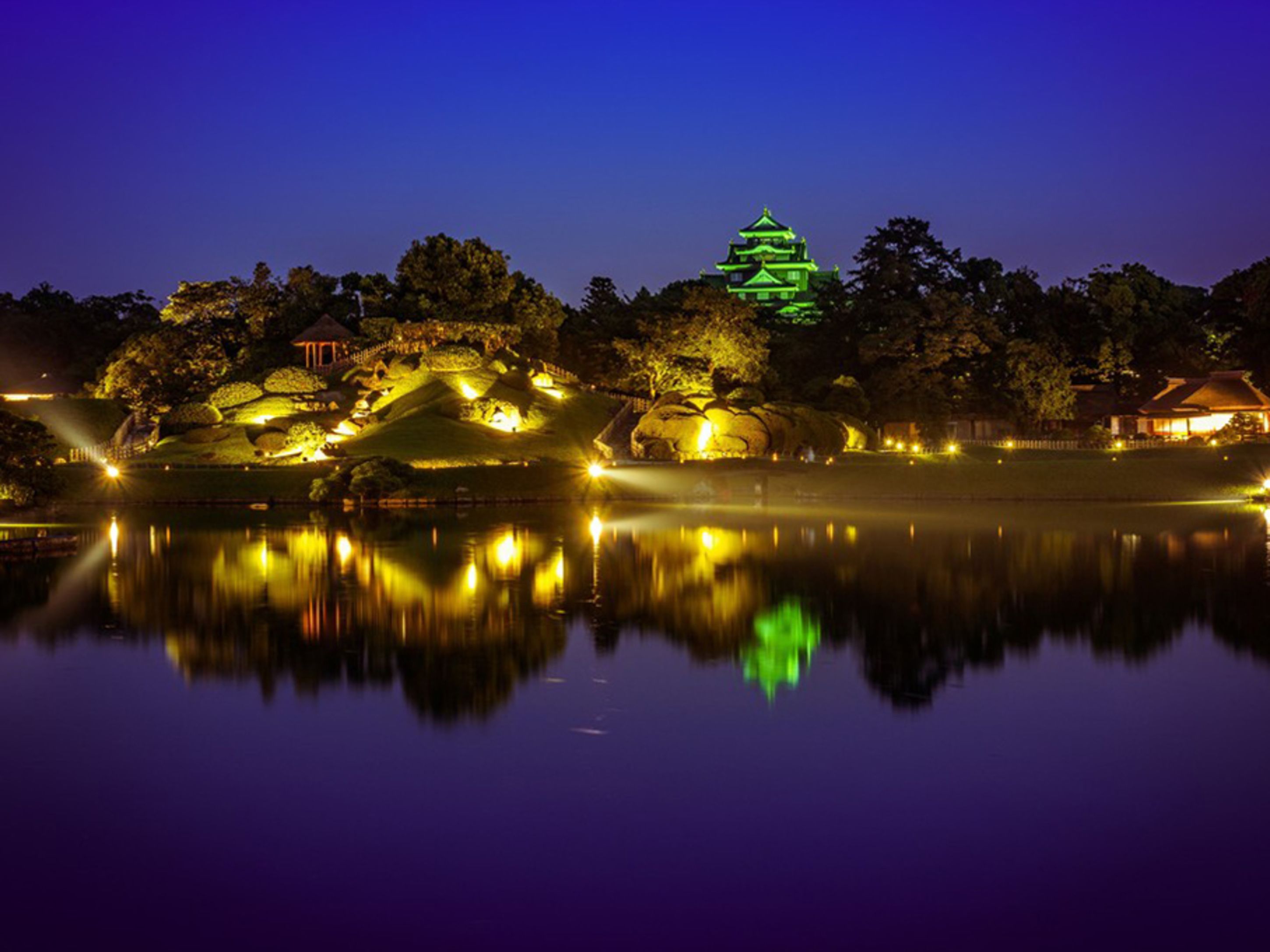 Un château japonais traditionnel, dont le reflet se mire dans l’eau, entouré d’arbres et illuminé la nuit.