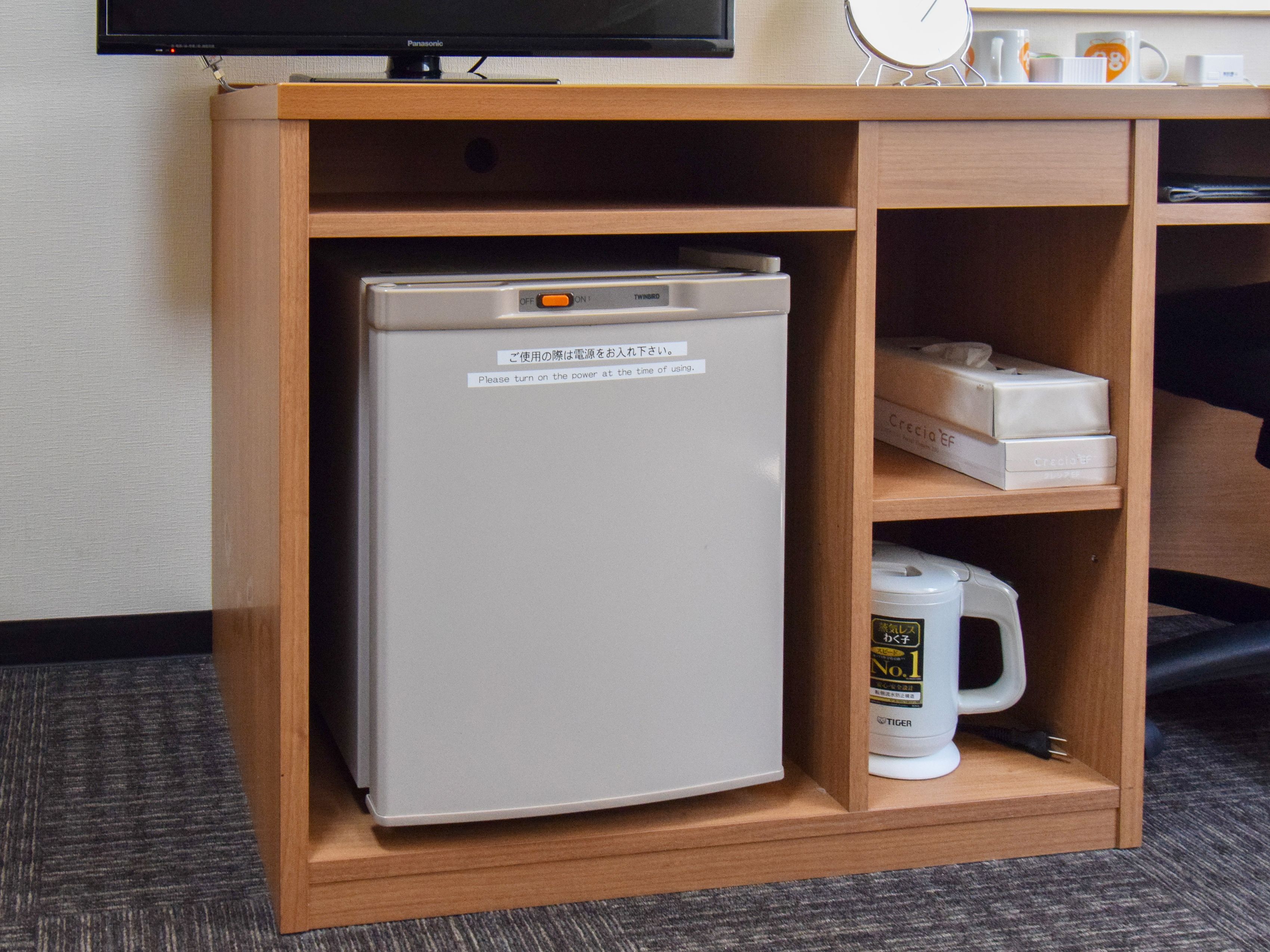 A compact refrigerator with a power switch, a coffee maker, and two boxes labeled "Crecia" in a wooden cabinet.
