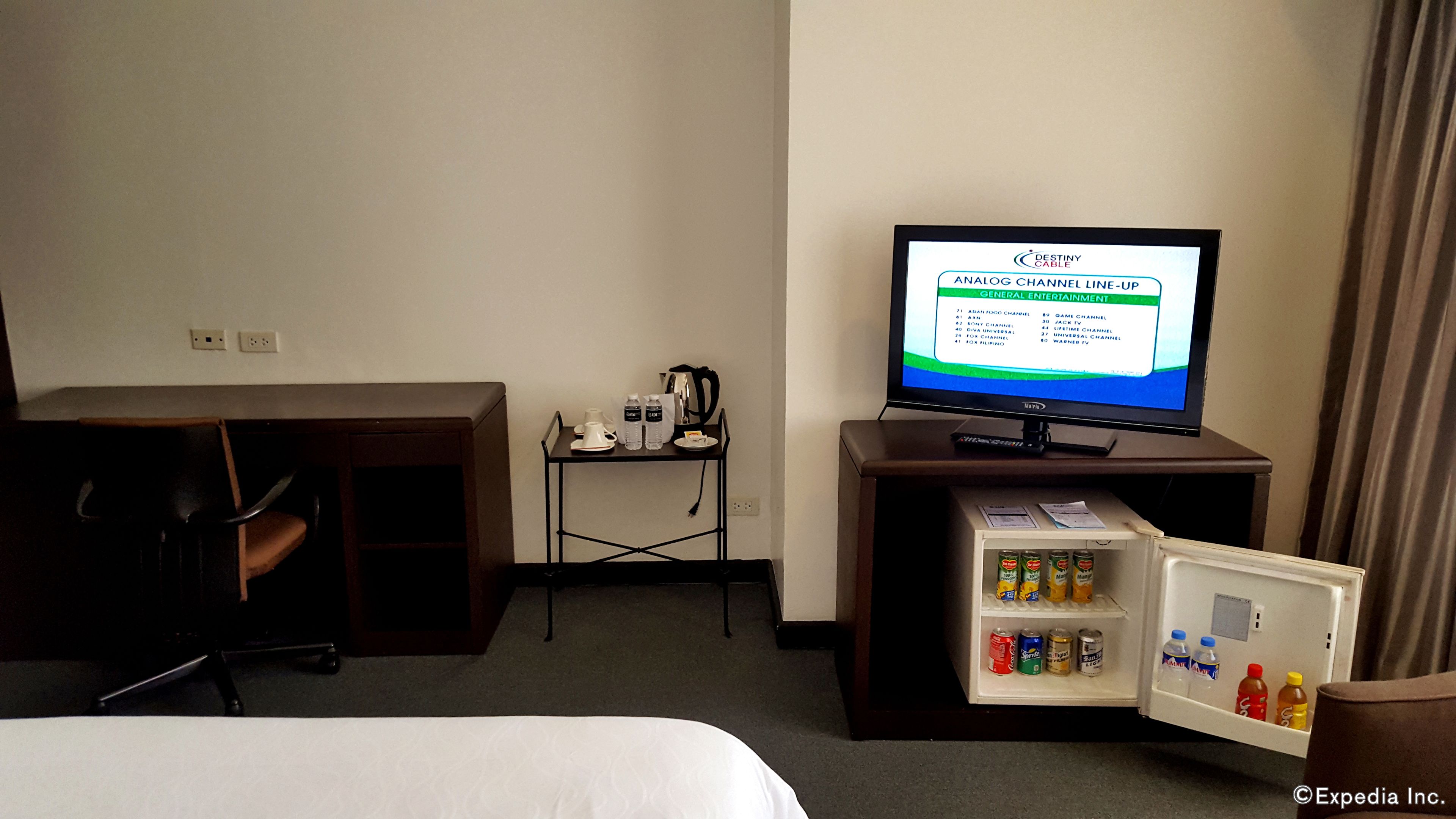 Quarto de hotel com cama, escrivaninha com computador, televisão exibindo lista de canais e mini-geladeira com bebidas.
