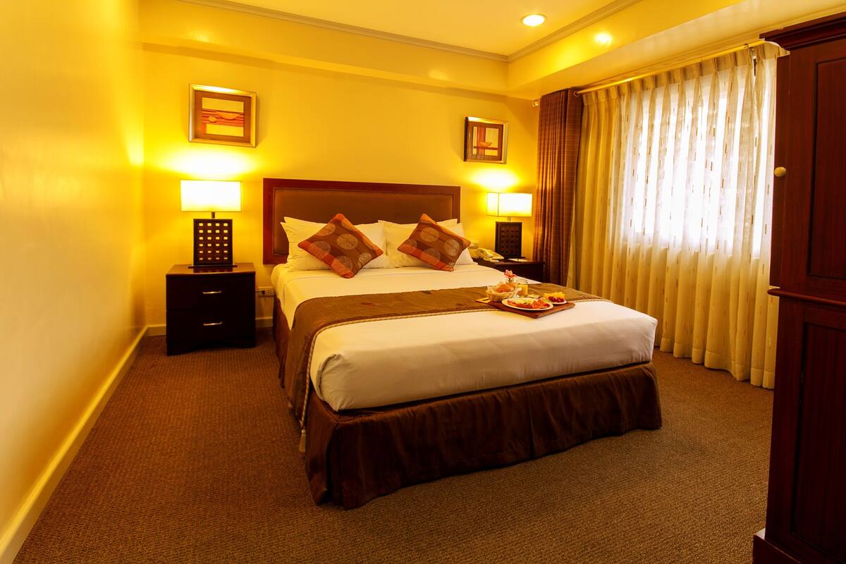 Habitación de hotel con una cama grande, dos lámparas de noche, un cabecero de madera y una ventana con cortinas.