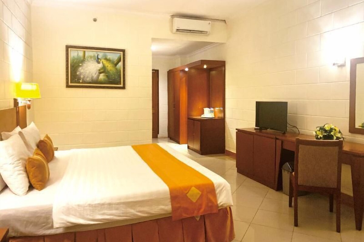 A hotel room with a bed, a desk, a chair, a television, a painting on the wall, and an air conditioning unit.