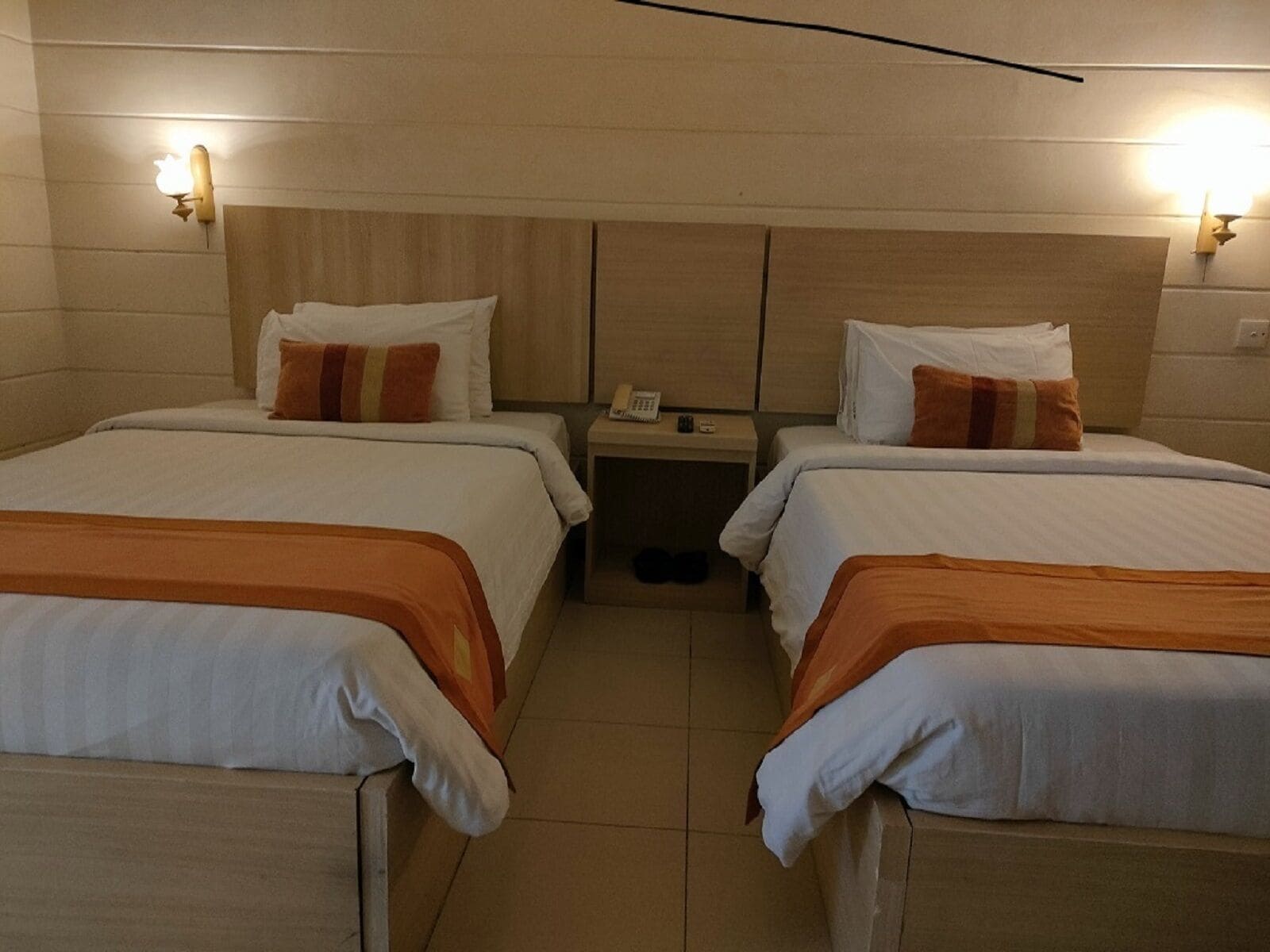A hotel room with two beds, each with white bedding and orange accents. There is a nightstand between the beds with a phone and a small lamp. The room has wooden headboards and walls.