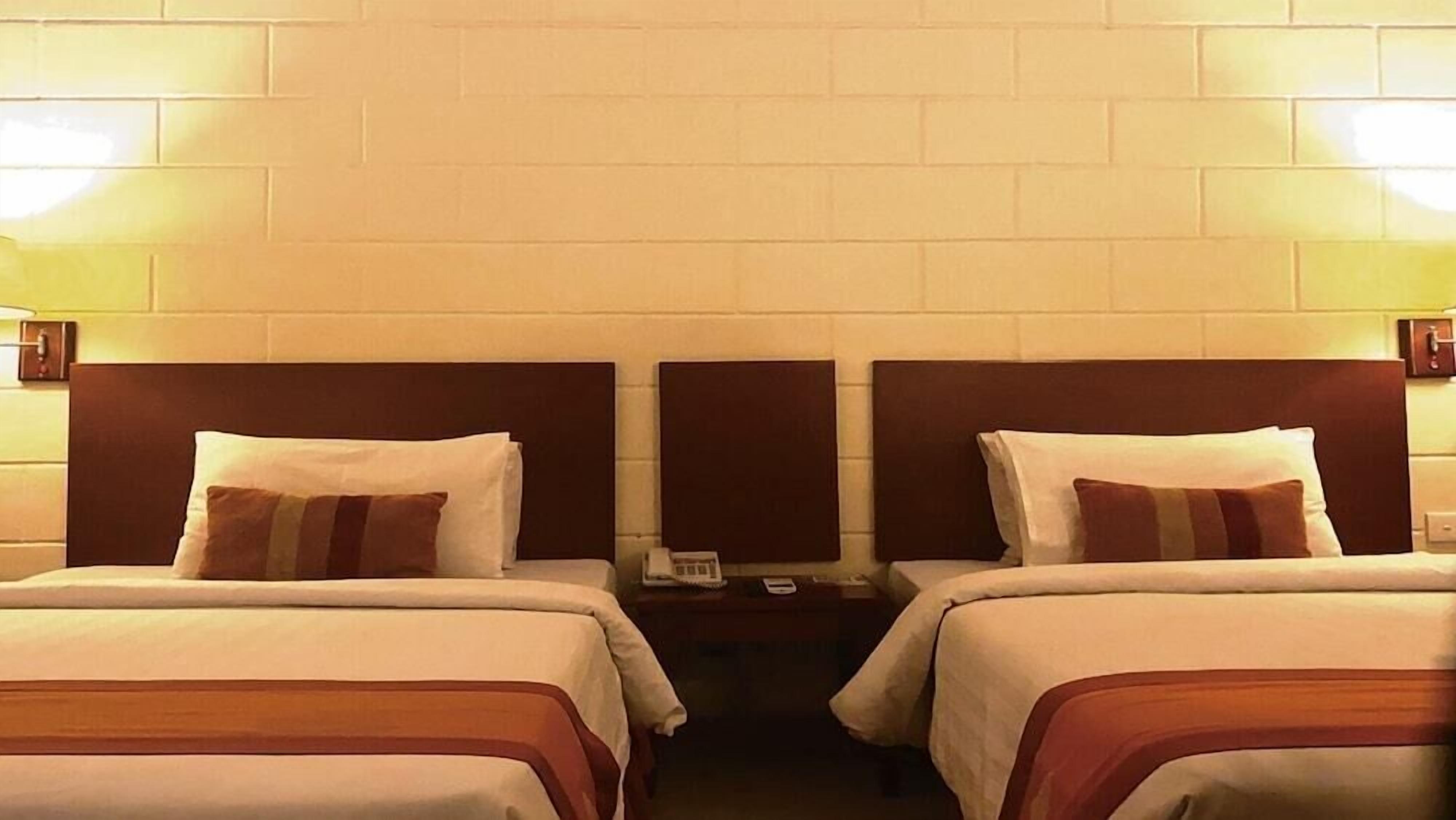 A hotel room with two beds, each with a headboard, a bedside table with a lamp, and a wall-mounted phone.