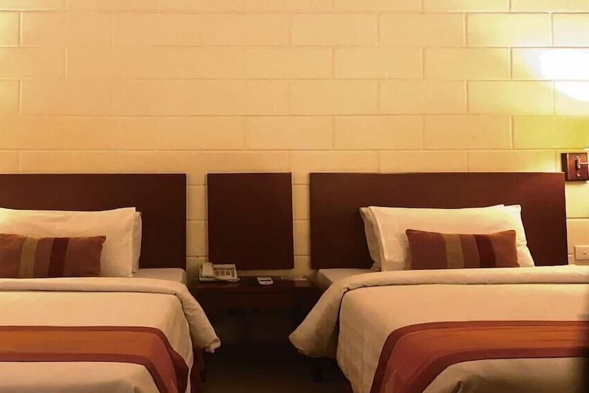 Una habitación de hotel con dos camas, cada una con cabecero, mesita de noche con lámpara y teléfono en la pared.