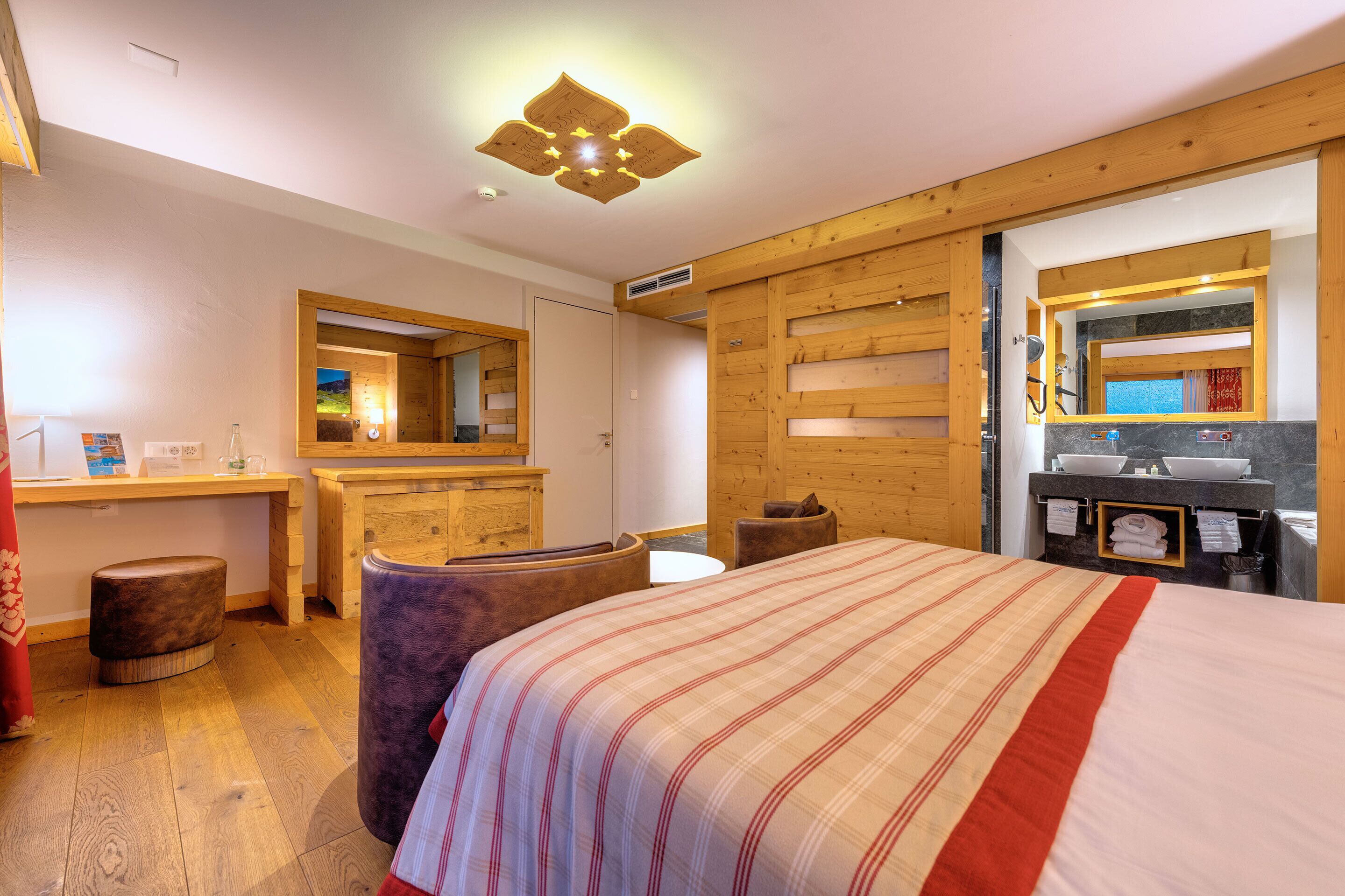 Ett hotellrum med en säng, ett skrivbord i trä, en spegel och ett badrum med handfat och handduksställ.