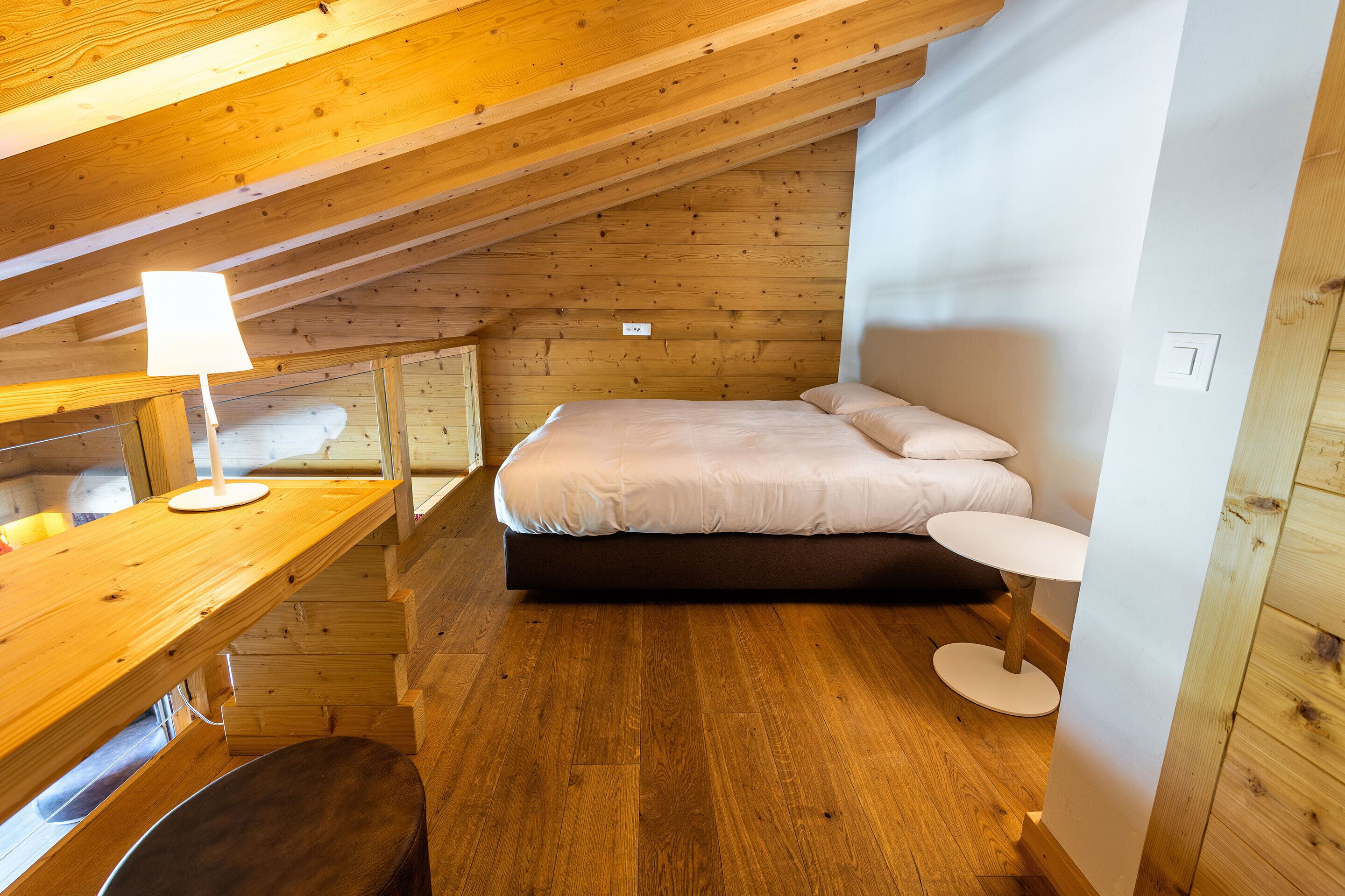Una habitació petita i acollidora amb un sostre de fusta, un llit amb roba de llit blanca, una taula rodona i un escriptori amb una làmpada.