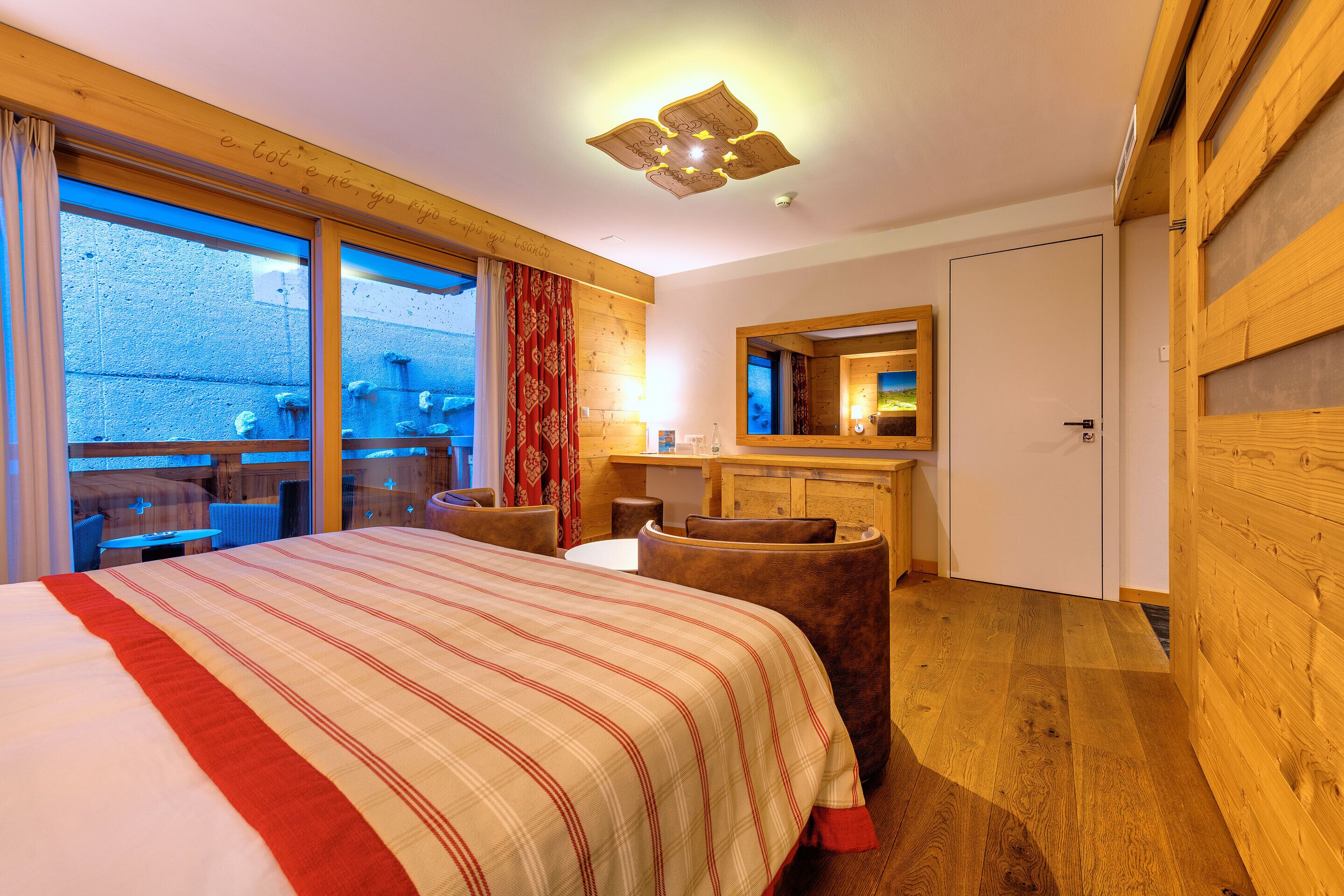Habitación de hotel con una cama grande, paredes de madera, techo de madera, un escritorio pequeño y un ventanal amplio.